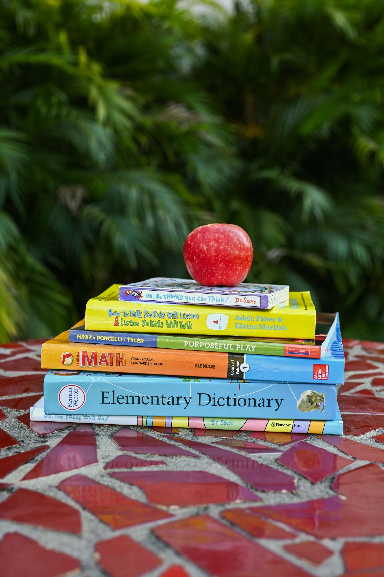 Stack of educational books with red apple on top against lush green background.
