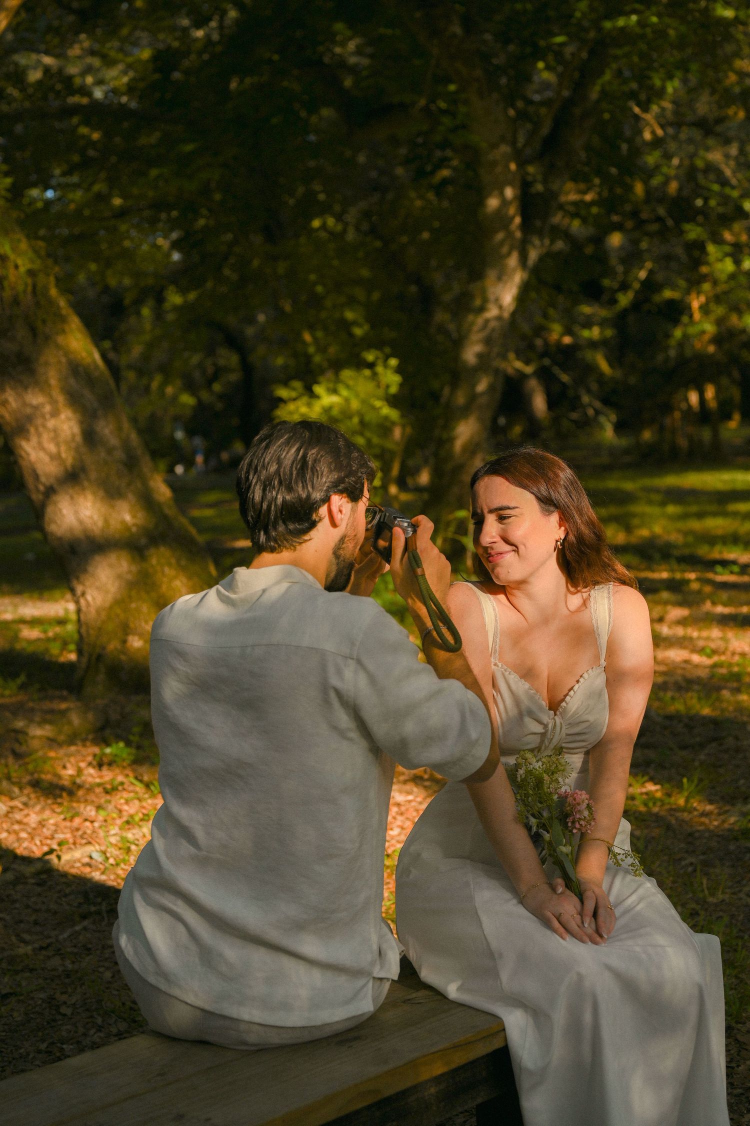 A couple shares a playful moment on a park bench surrounded by golden sunlit trees.