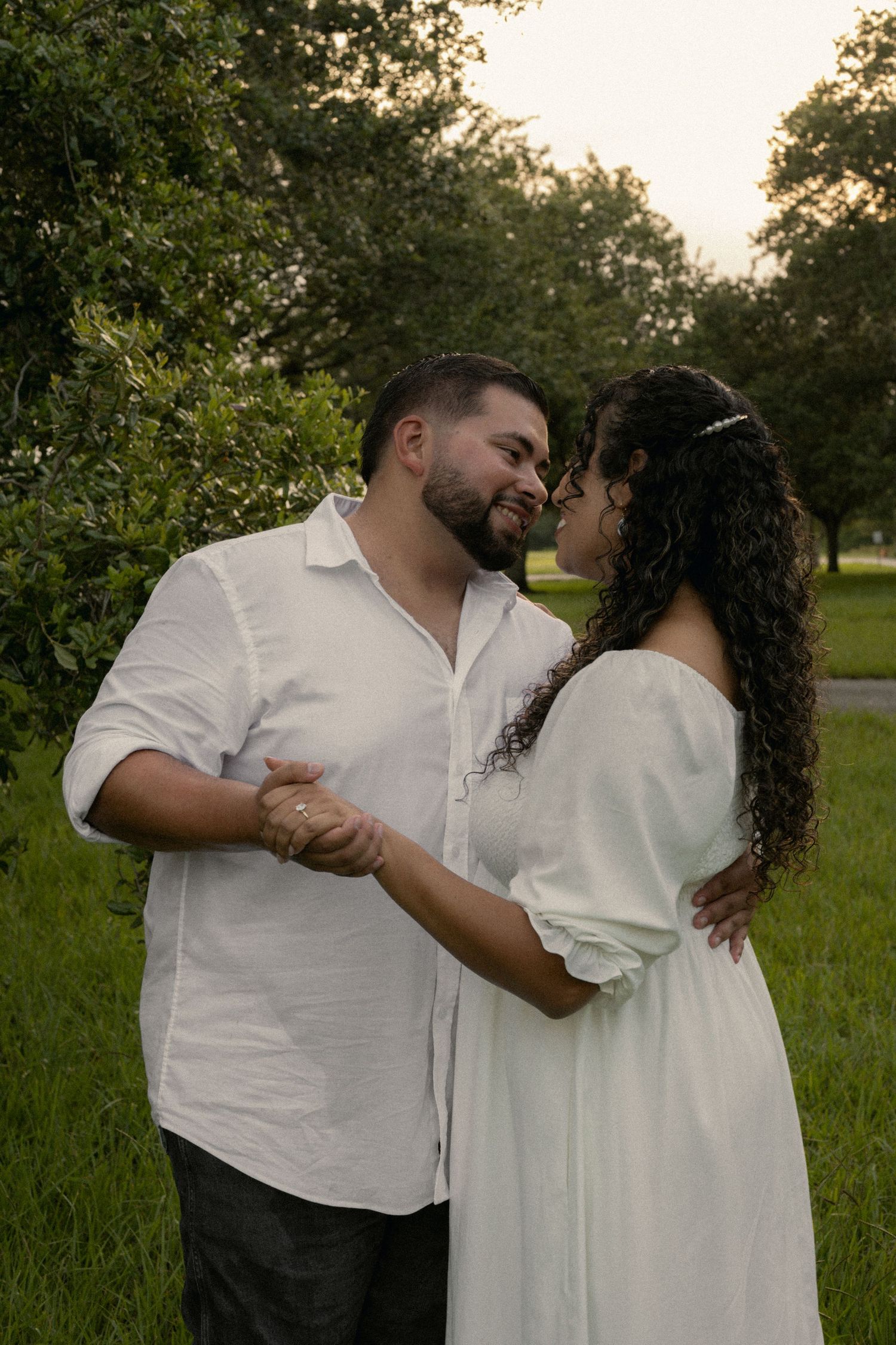 Couple in white outfits sharing a romantic dance outdoors, surrounded by lush green trees at dusk.