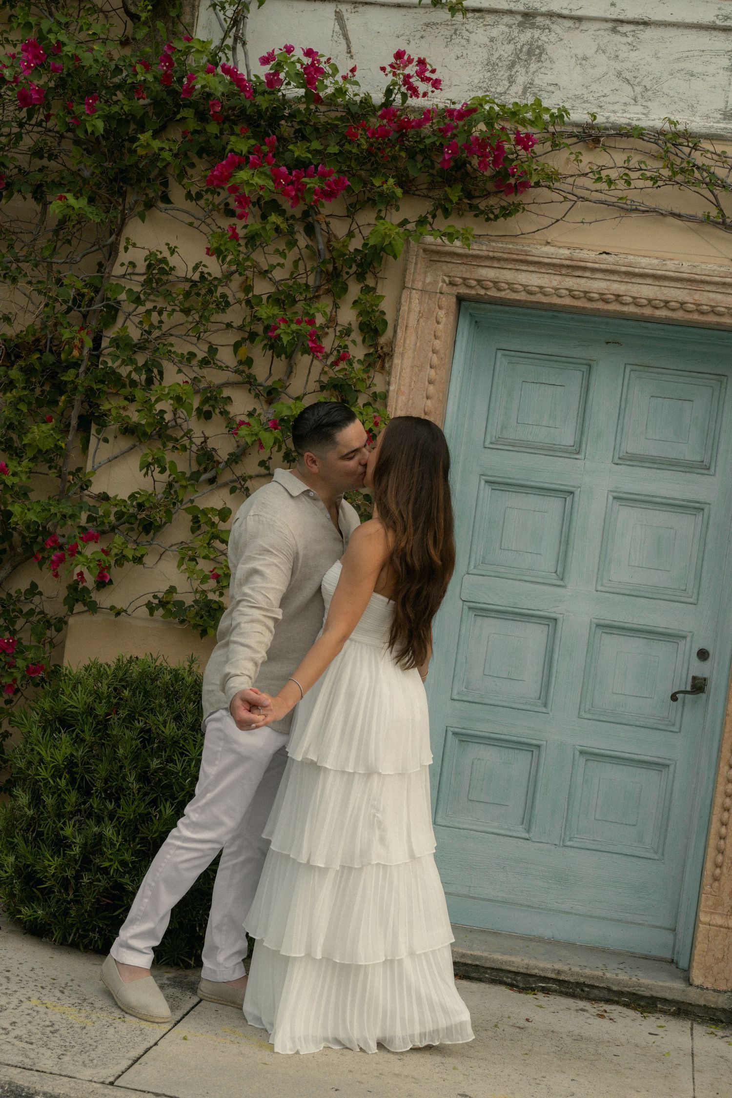 Couple sharing a romantic kiss in white outfits by a teal door with pink bougainvillea flowers on a wall.