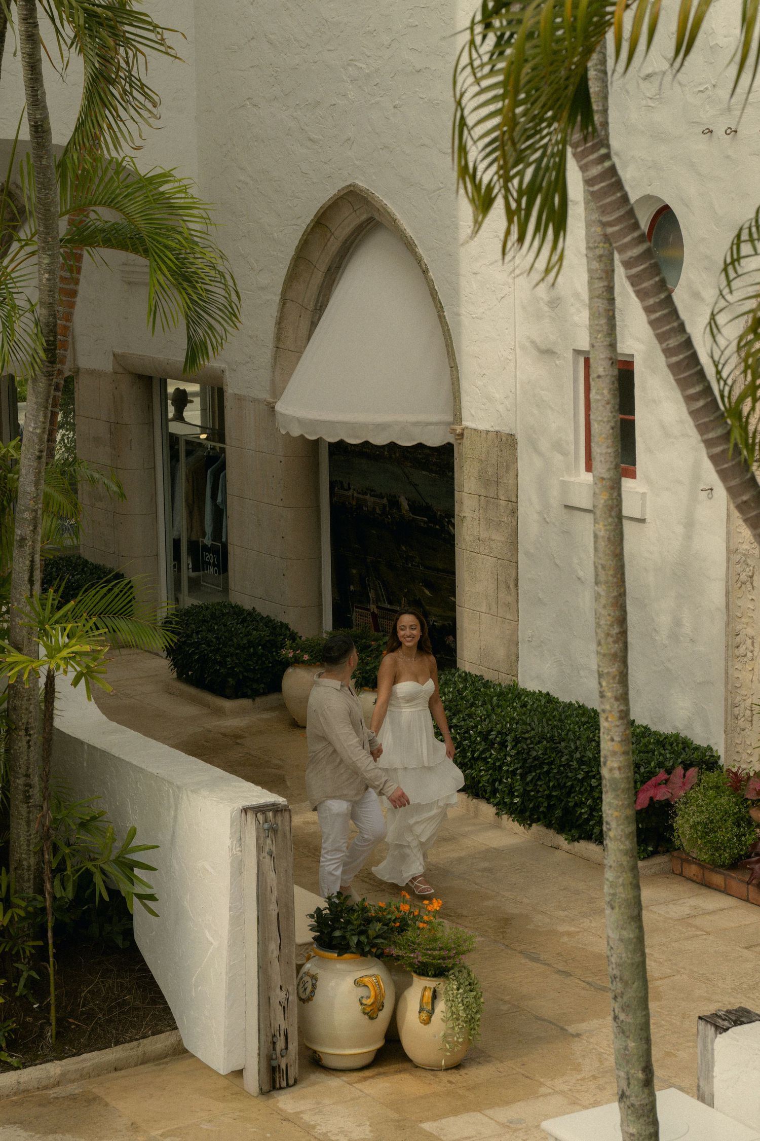 Couple in white attire holding hands near tropical resort entrance with palm trees and arched doorway.