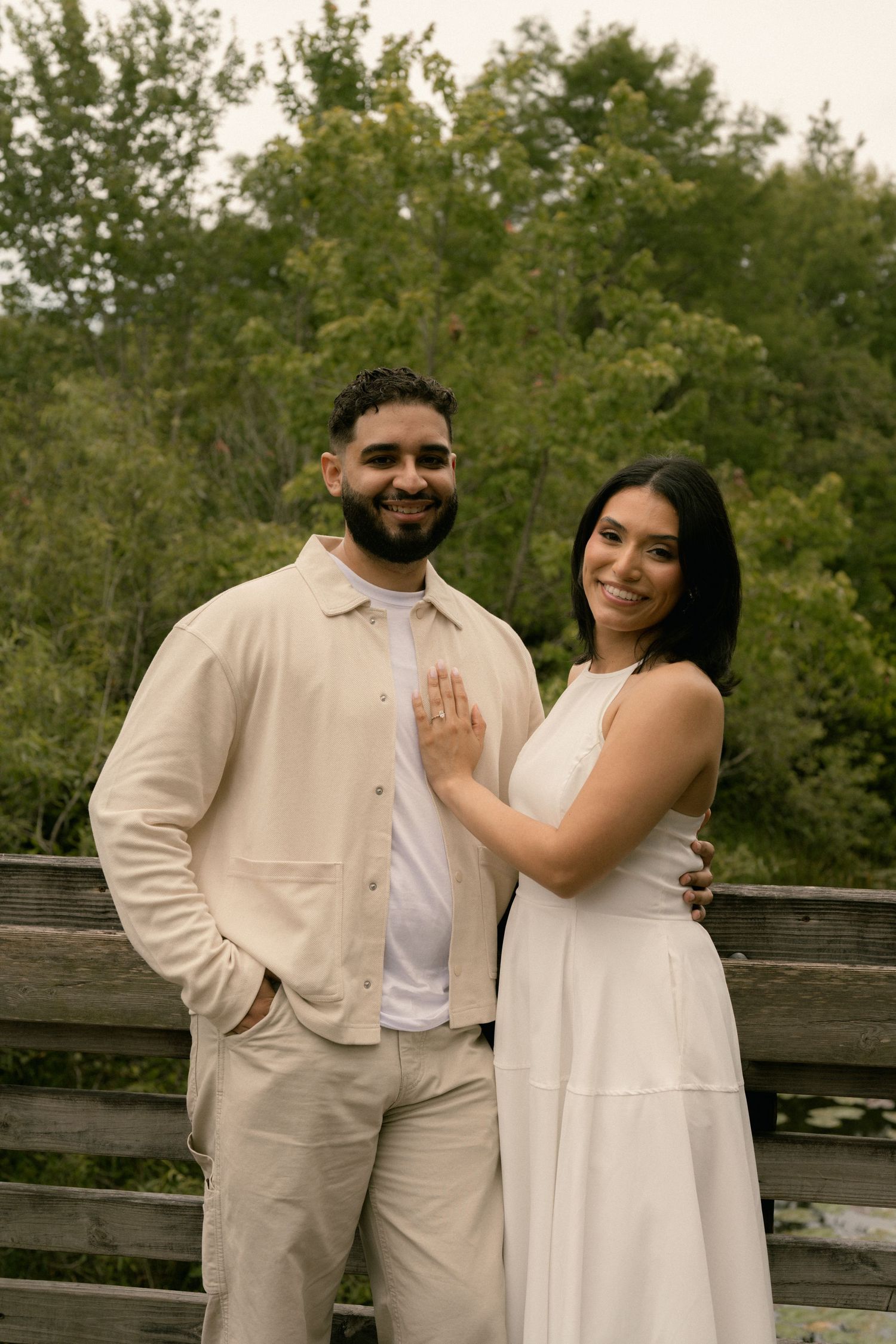 Engaged couple poses on a wooden bridge smiling, woman showing off her engagement ring.