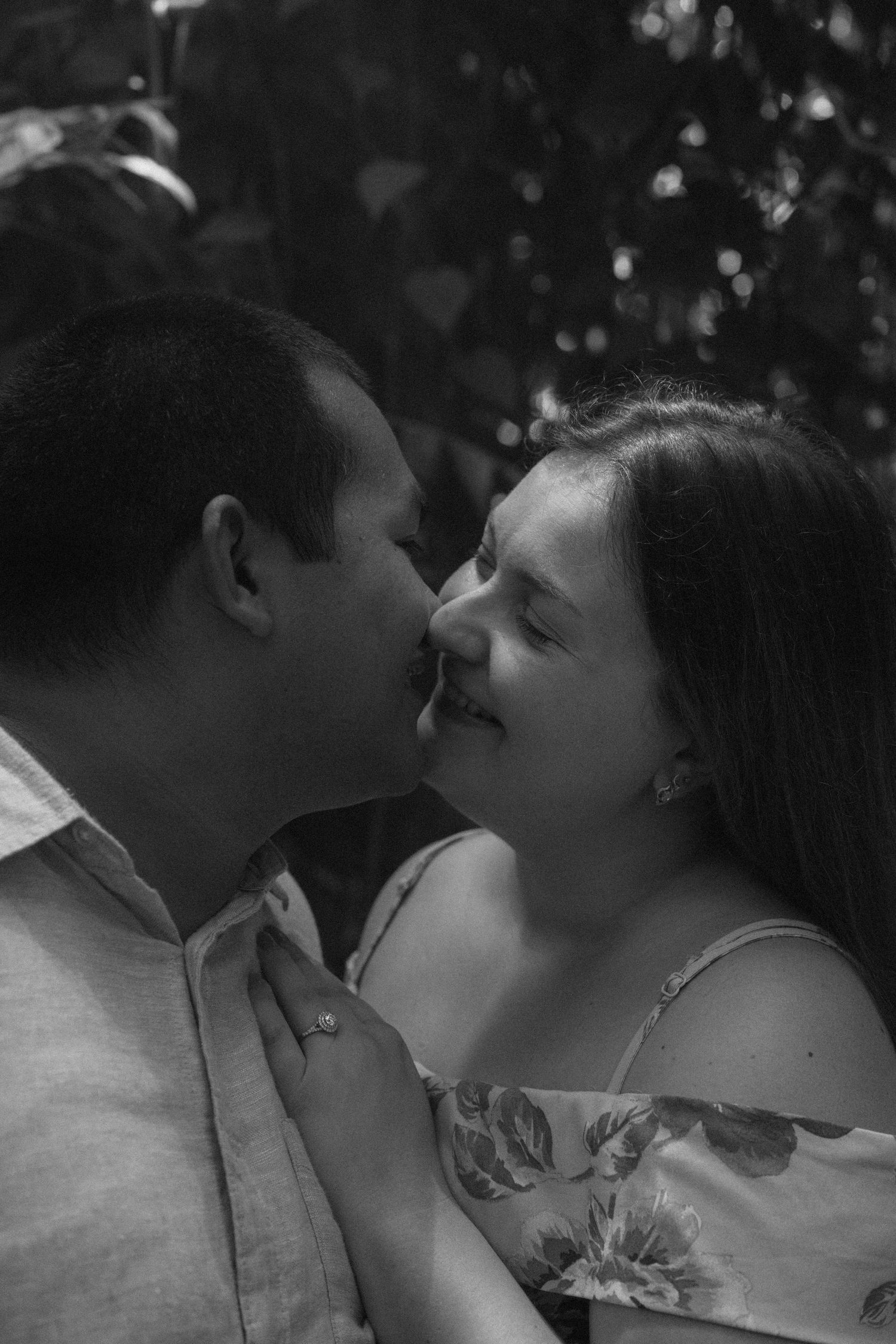 Black and white photo of a couple sharing a romantic moment, almost kissing, surrounded by lush greenery.