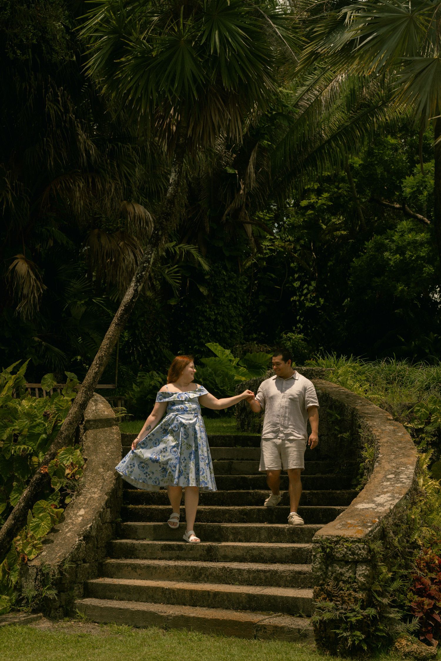 Couple holding hands on curved stone steps surrounded by lush tropical palm trees and greenery.
