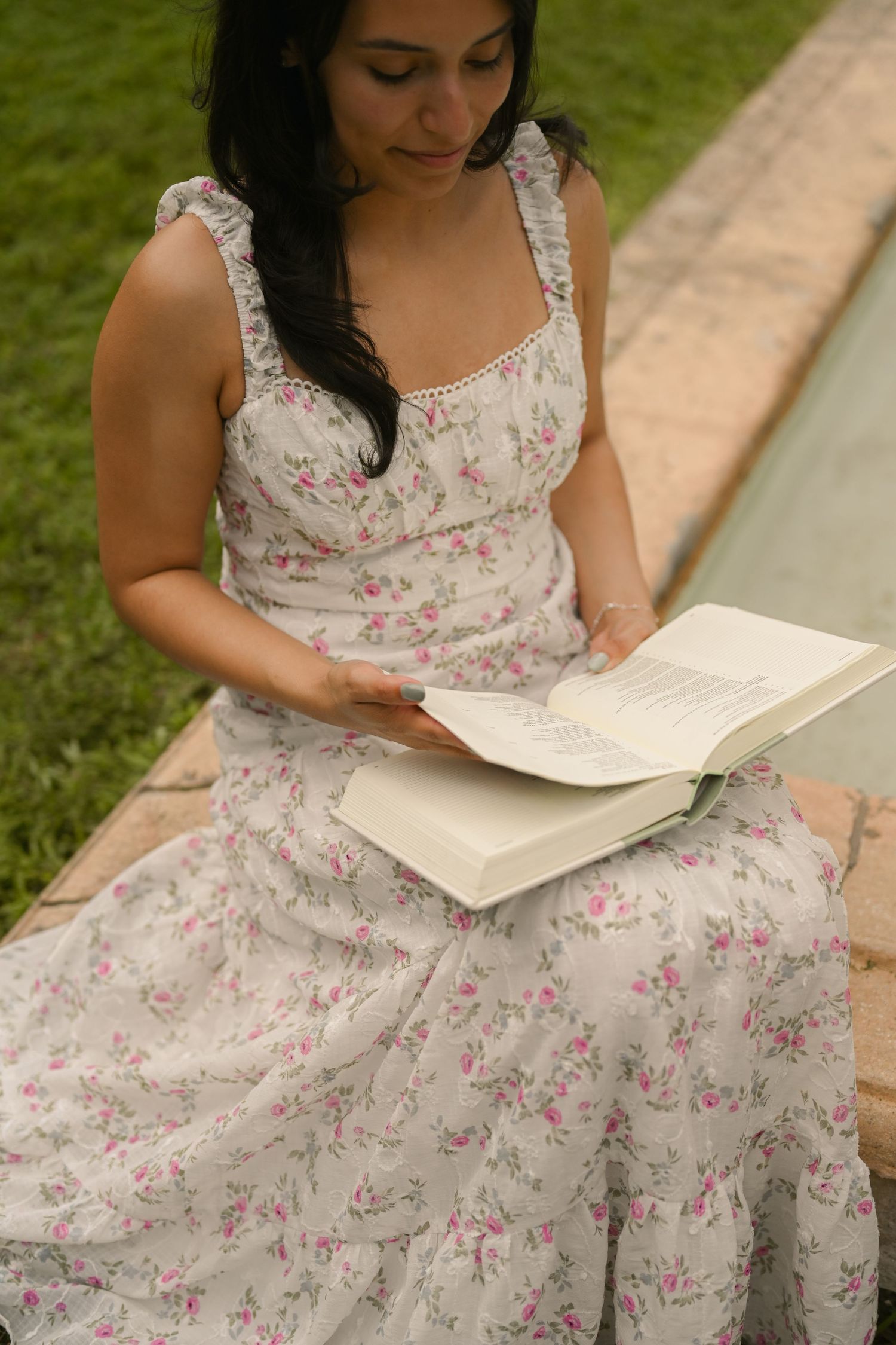 A person in a floral dress sits on grass while reading a book in a peaceful outdoor setting.