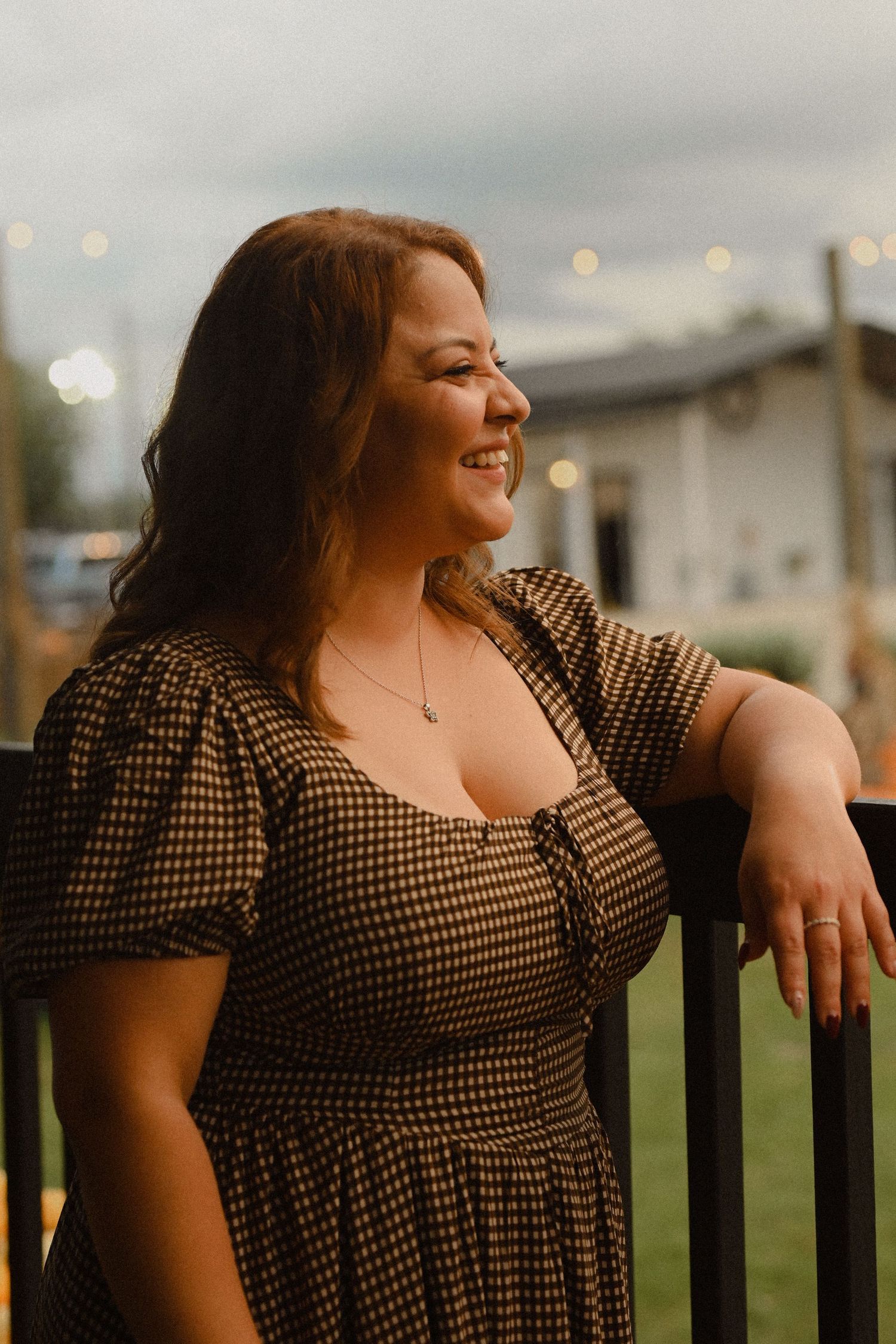 A person in a polka dot dress smiles and leans on a railing outdoors at dusk with string lights in the background.