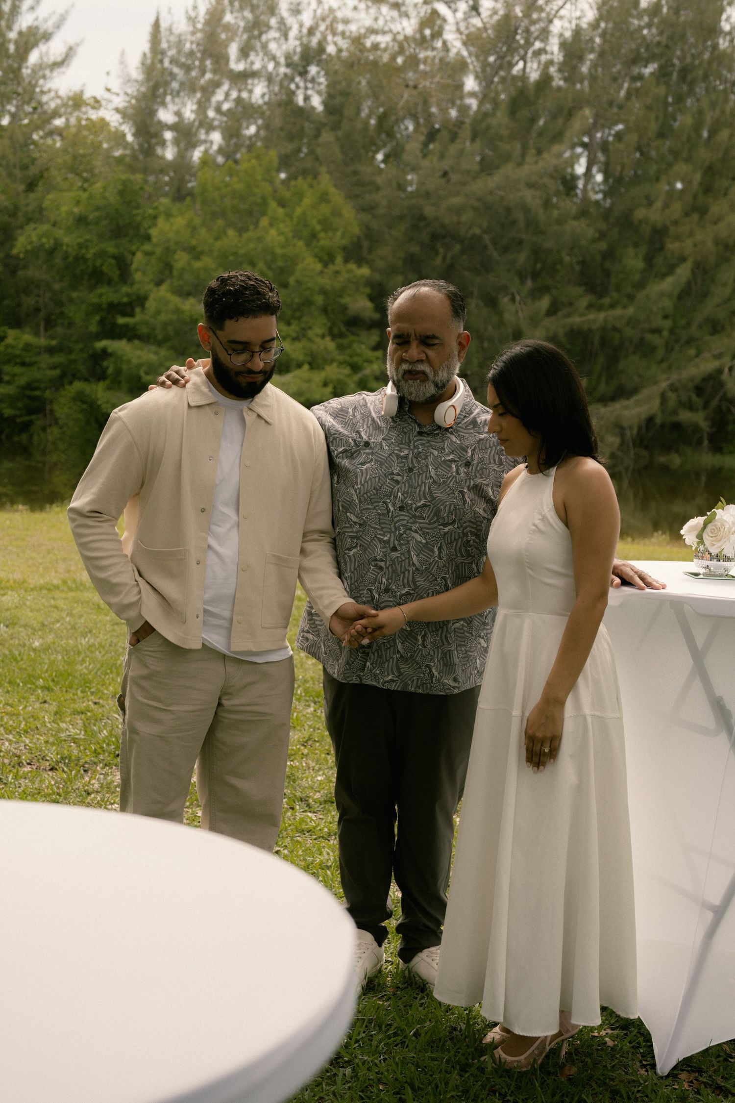 Three people gathered outdoors at a garden event, two men and a woman in white dress near a table.