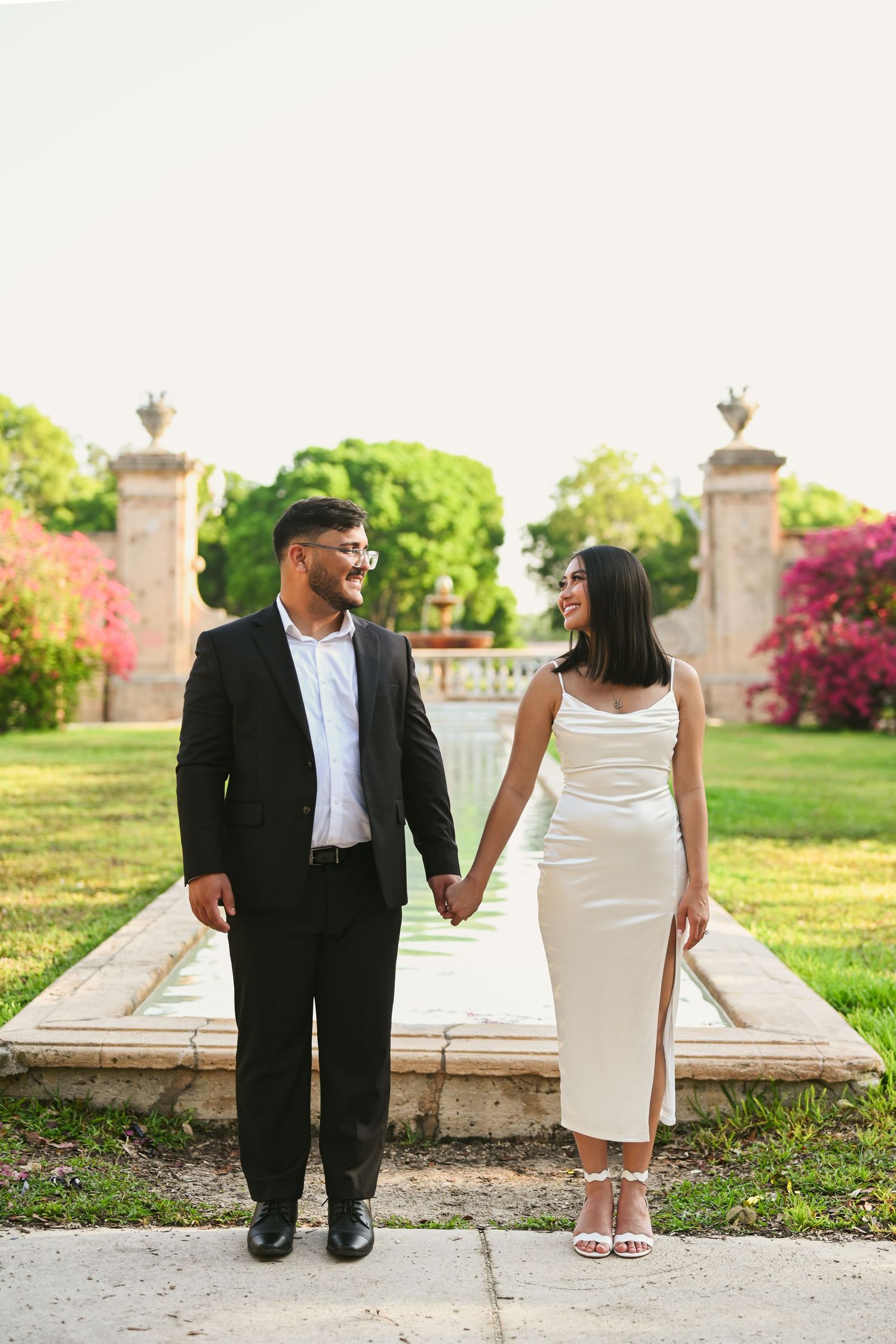 Couple in formal attire posing together on stone steps surrounded by lush garden scenery.