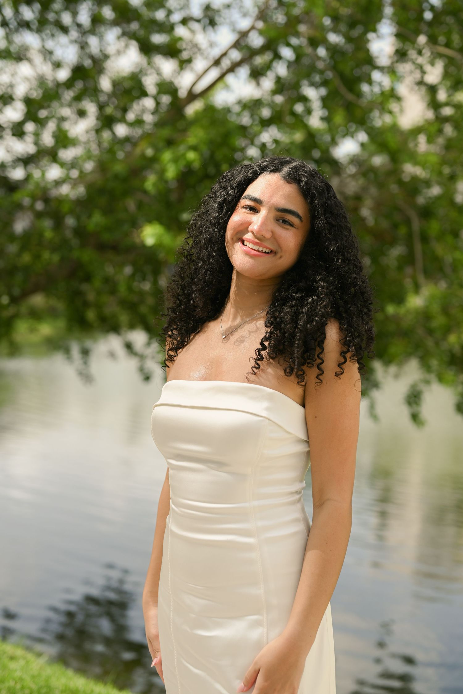 Graduate in strapless white dress poses against lakeside scenery during photo session.
