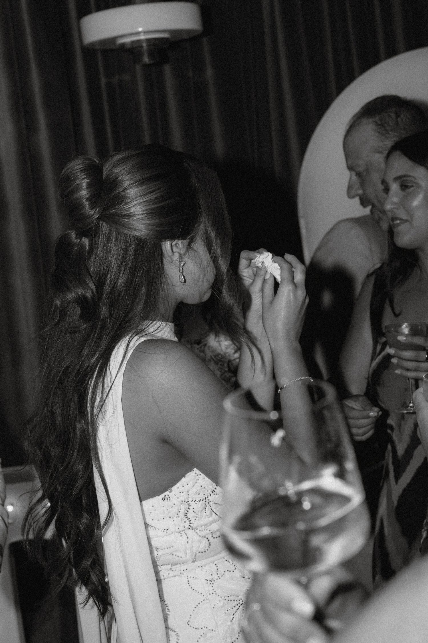 Black and white photo of a bride wiping tears at her wedding reception, surrounded by guests holding drinks.