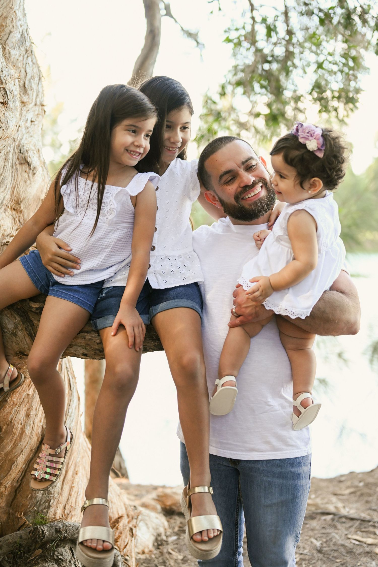 A smiling father holds his baby girl while two older daughters sit on a tree, all wearing white tops outdoors.