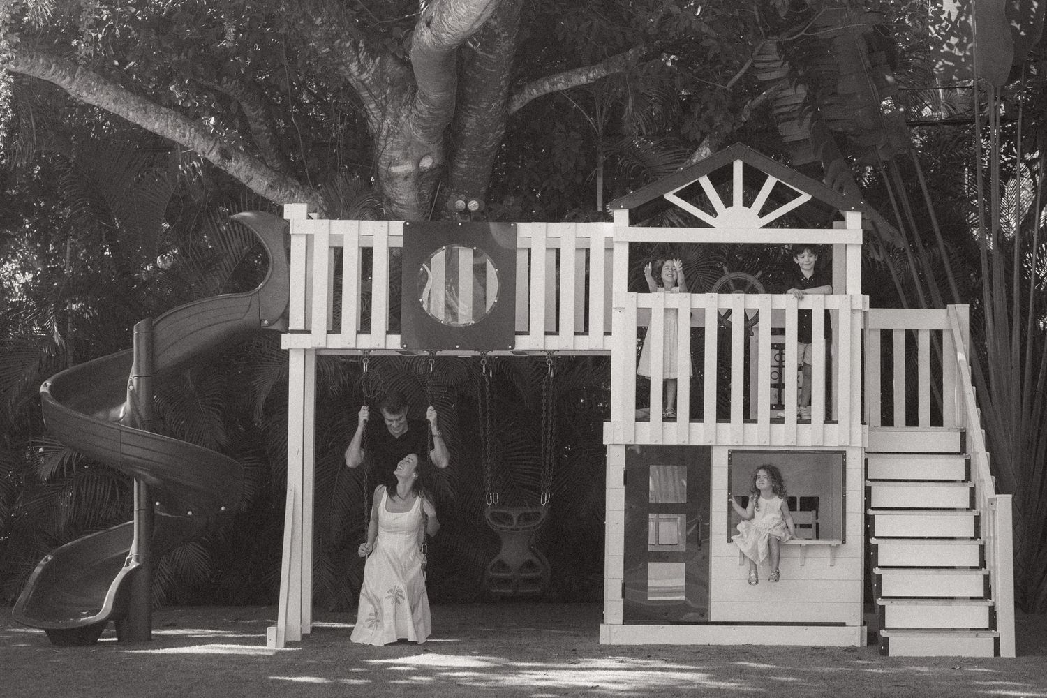 Black and white photo of a pregnant woman in a white dress standing by a wooden backyard playhouse with a spiral slide and children playing.