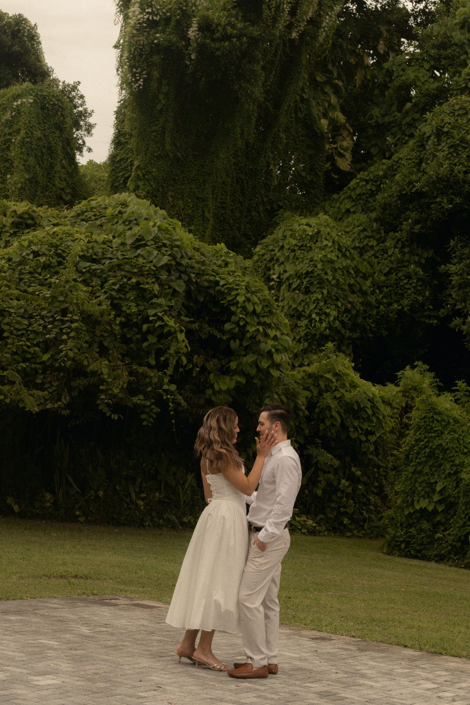 Couple in white embracing on a stone path in front of lush green hedges and trees, in a romantic engagement-style photo.