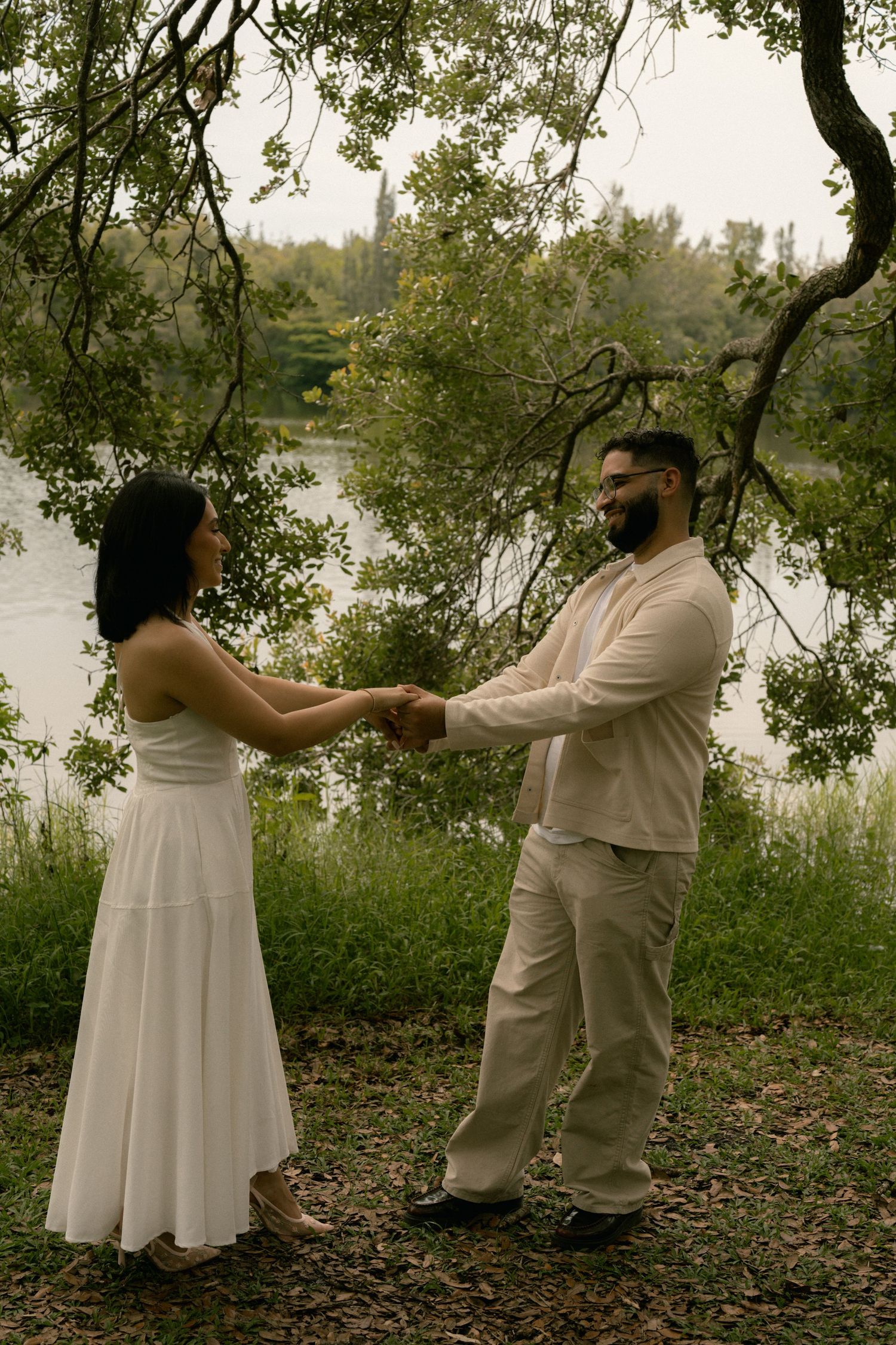 Couple holding hands in white outfits by a serene lake surrounded by lush green trees.