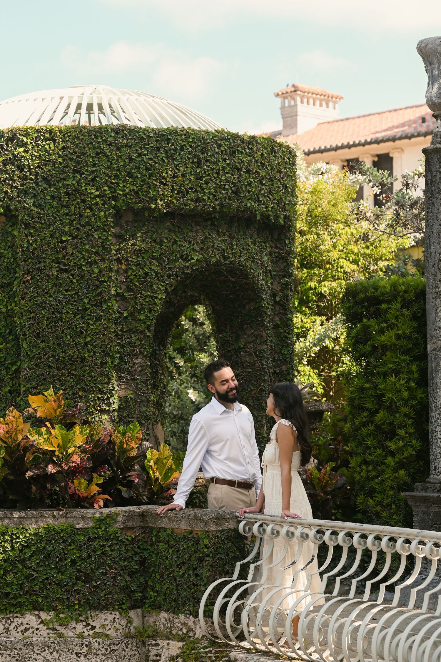 A couple shares an intimate moment on a Mediterranean-style balcony surrounded by manicured hedges.