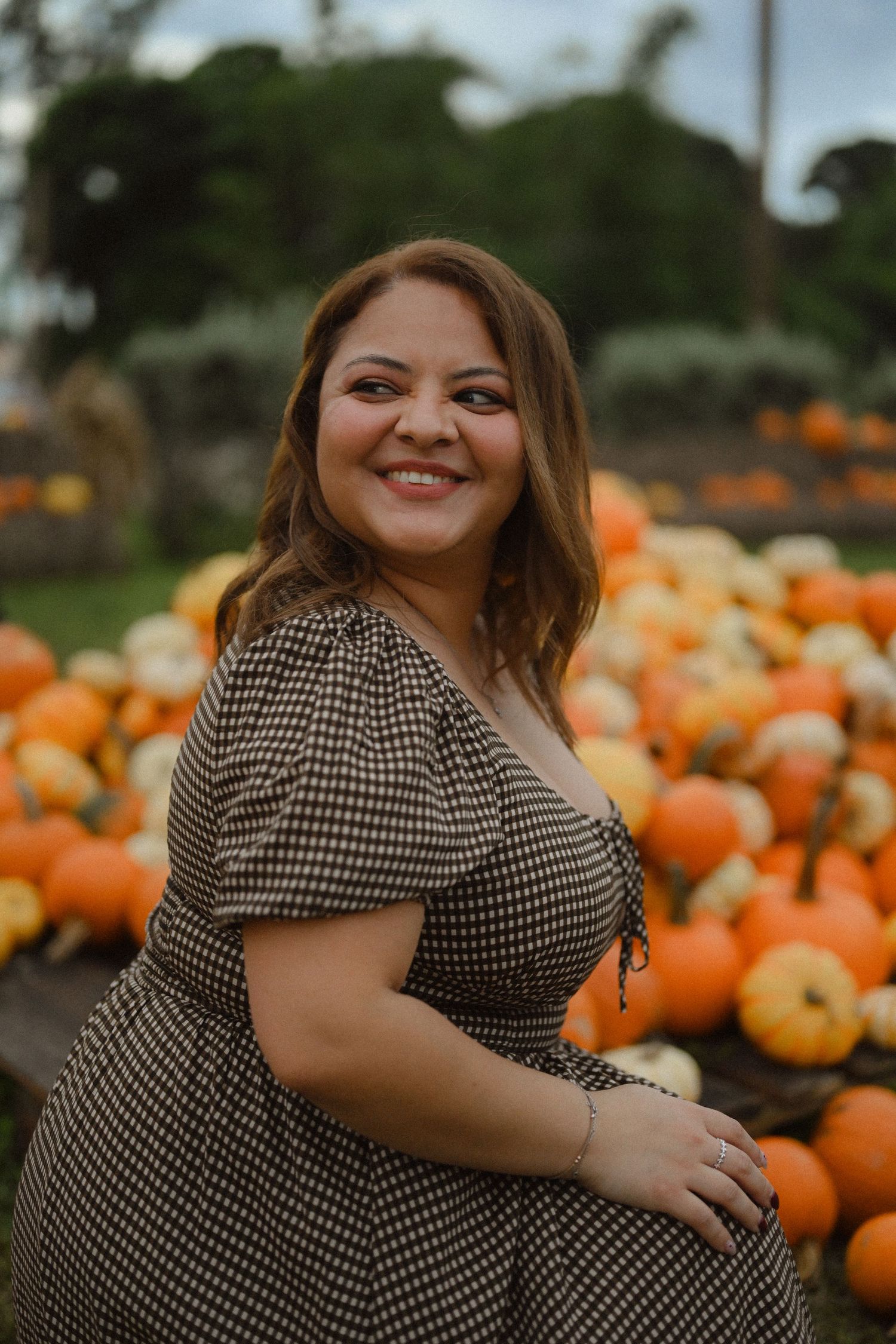 A person in a black and white checkered dress poses at a pumpkin patch surrounded by bright orange pumpkins.