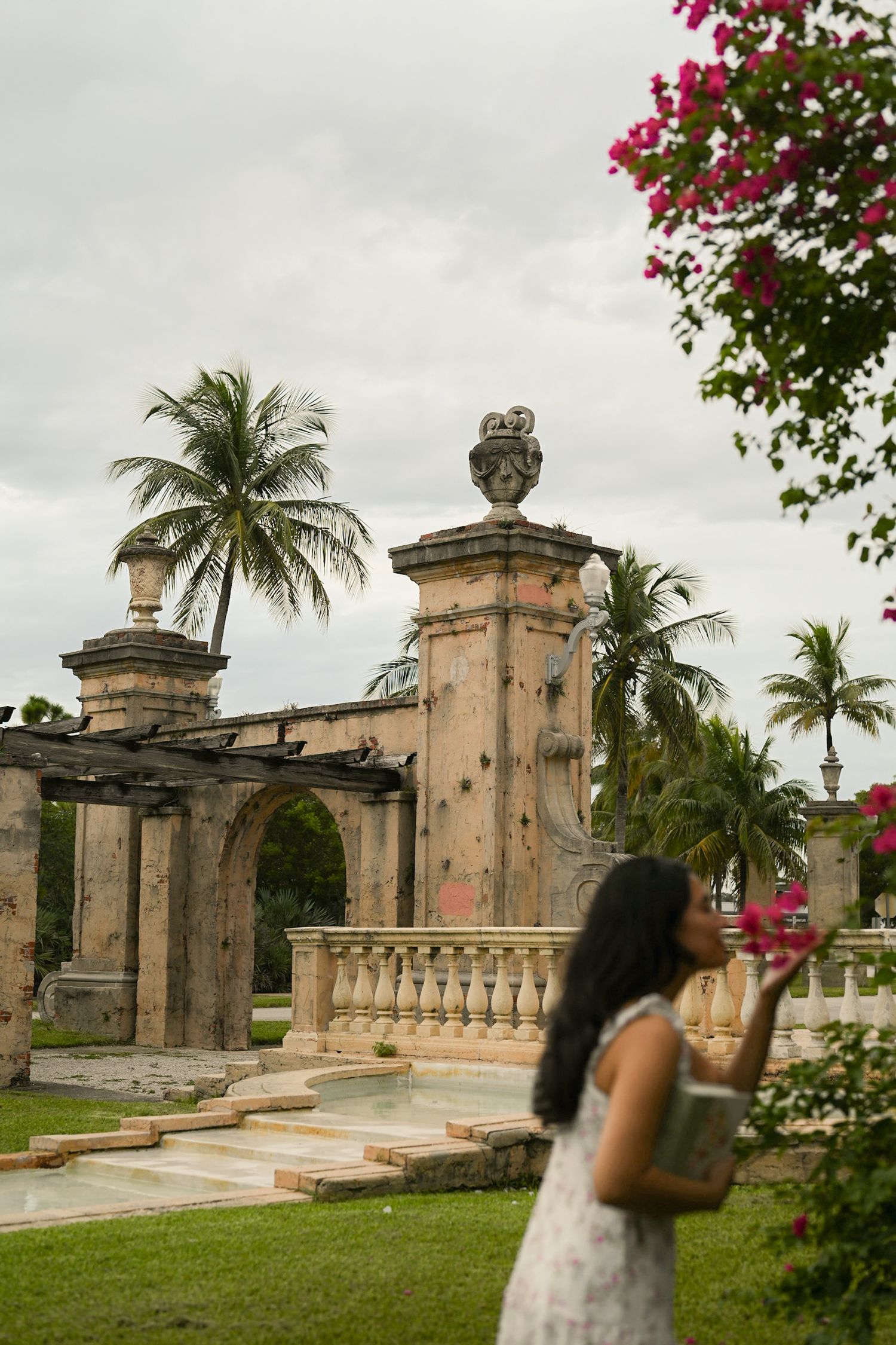 Historic stone pillars and palm trees frame a garden pathway under cloudy skies.