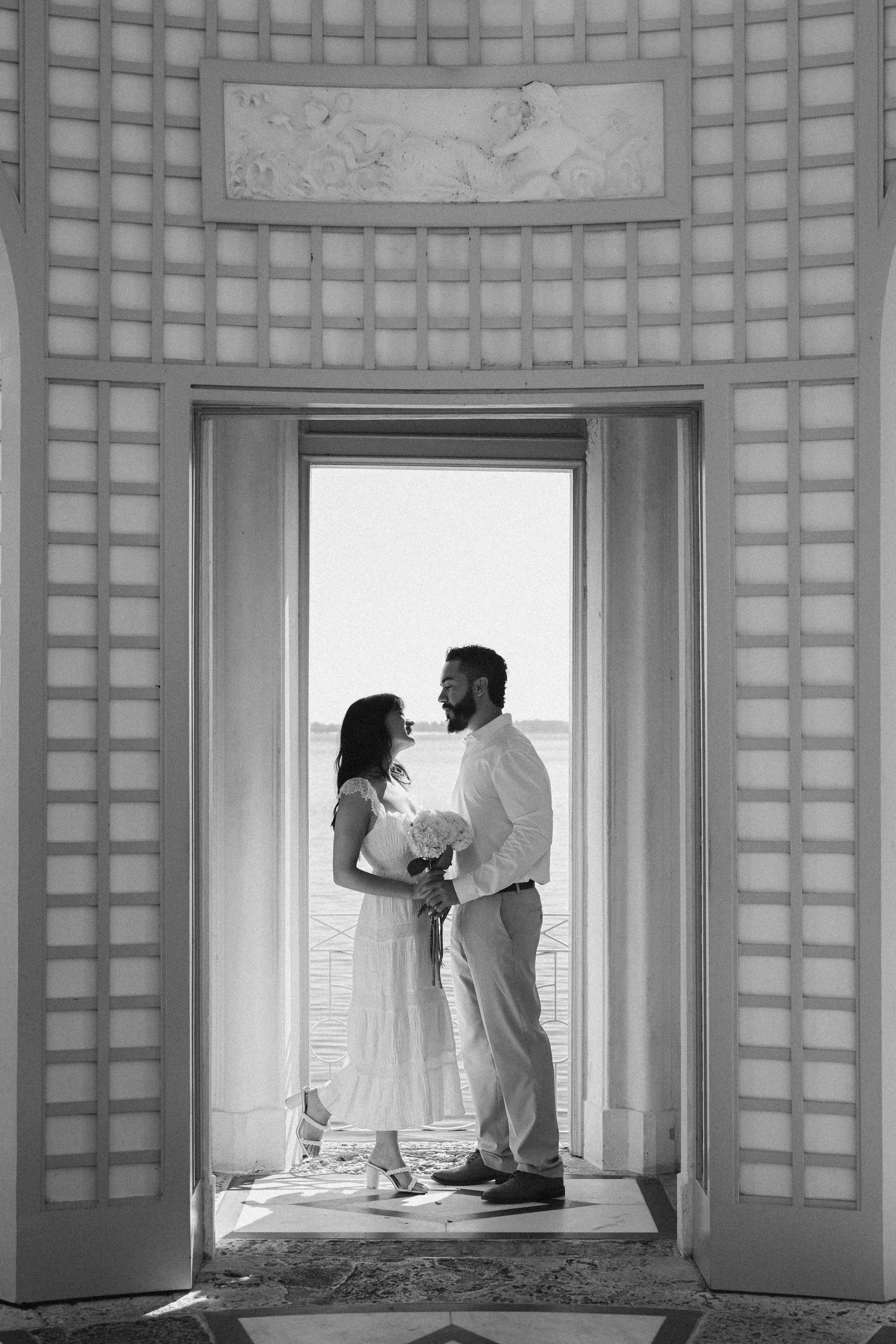 Black and white photo of a couple standing in an elegant doorway, sharing a romantic moment with ocean views behind them.