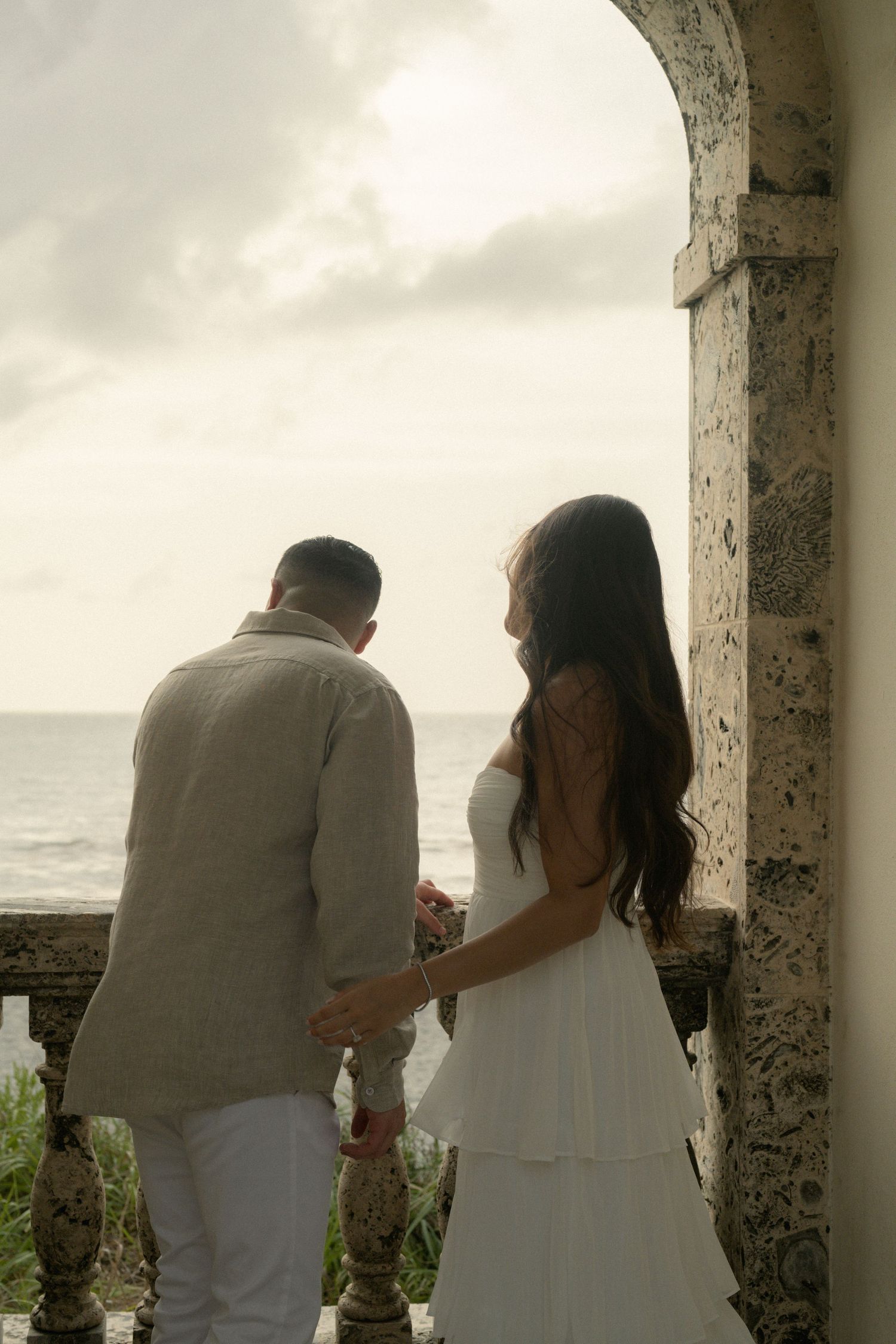 Couple in white attire standing on a stone balcony overlooking the ocean on a cloudy day.