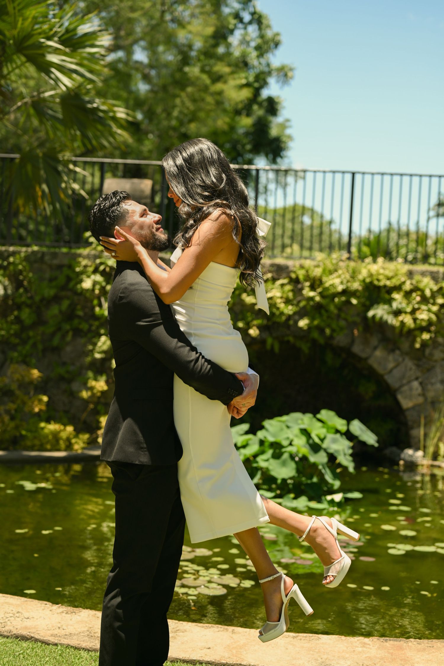 A couple in formal attire spins joyfully in a sunny garden setting.