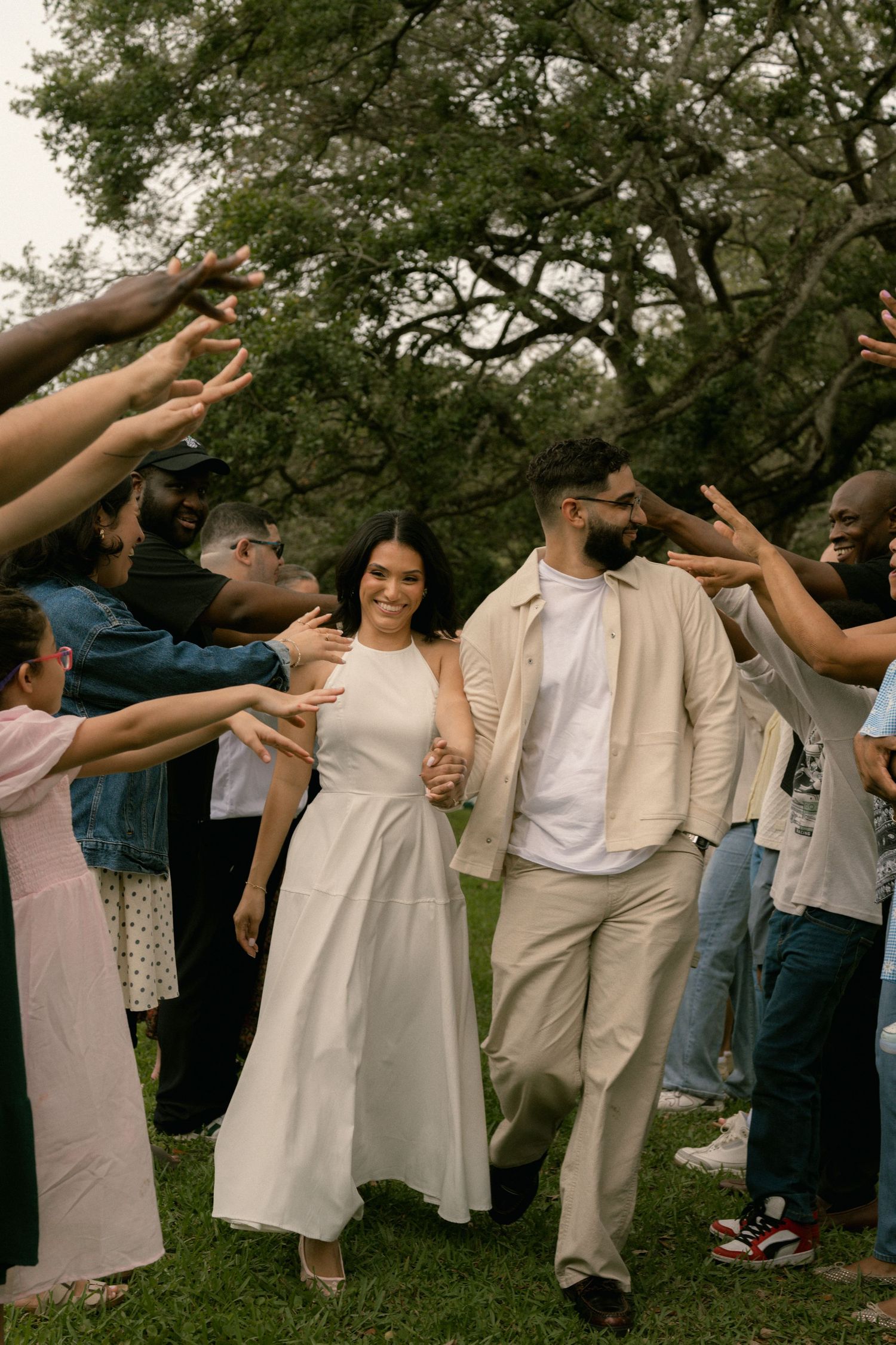 Joyful newlywed couple walks hand-in-hand through a celebratory tunnel of hands formed by cheering guests in a park.