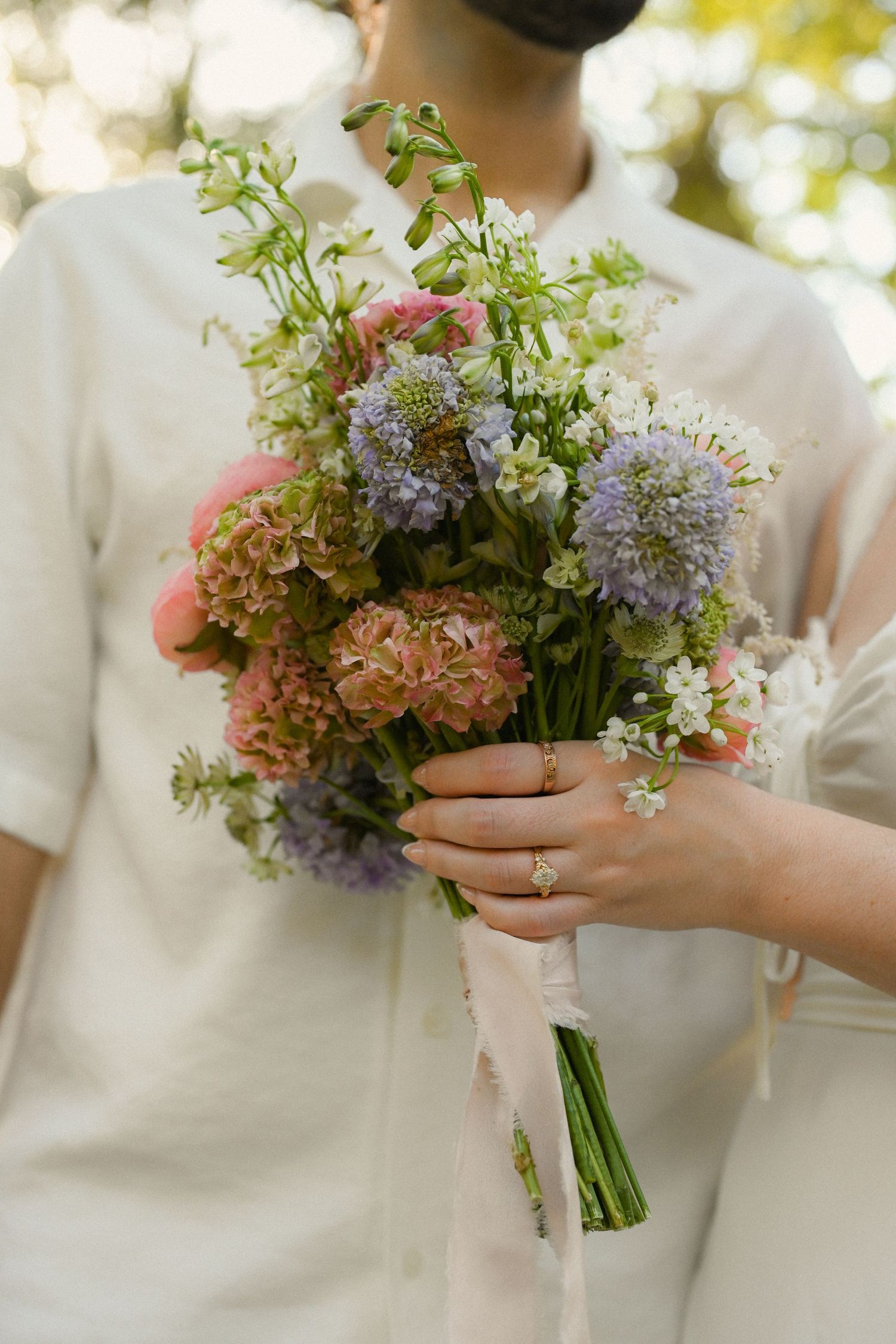 A delicate bouquet of pastel flowers including hydrangeas and alliums tied with ribbon is held against a white dress.