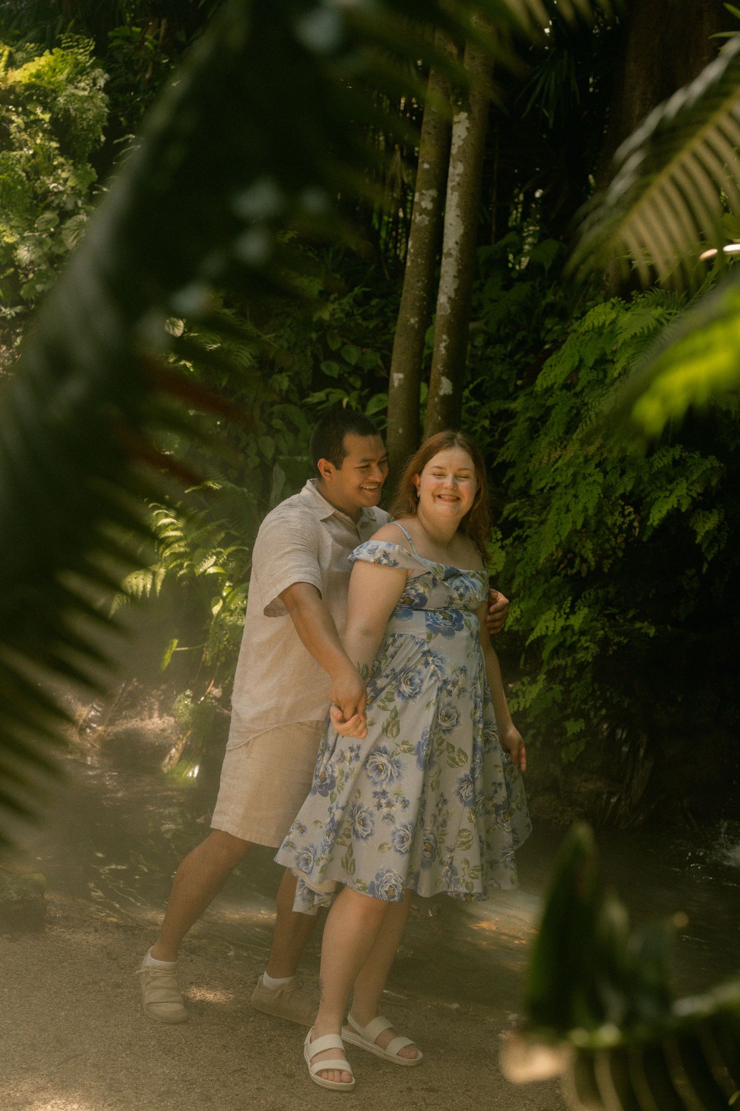 Couple holding hands and smiling on a sunlit garden path surrounded by lush tropical greenery.