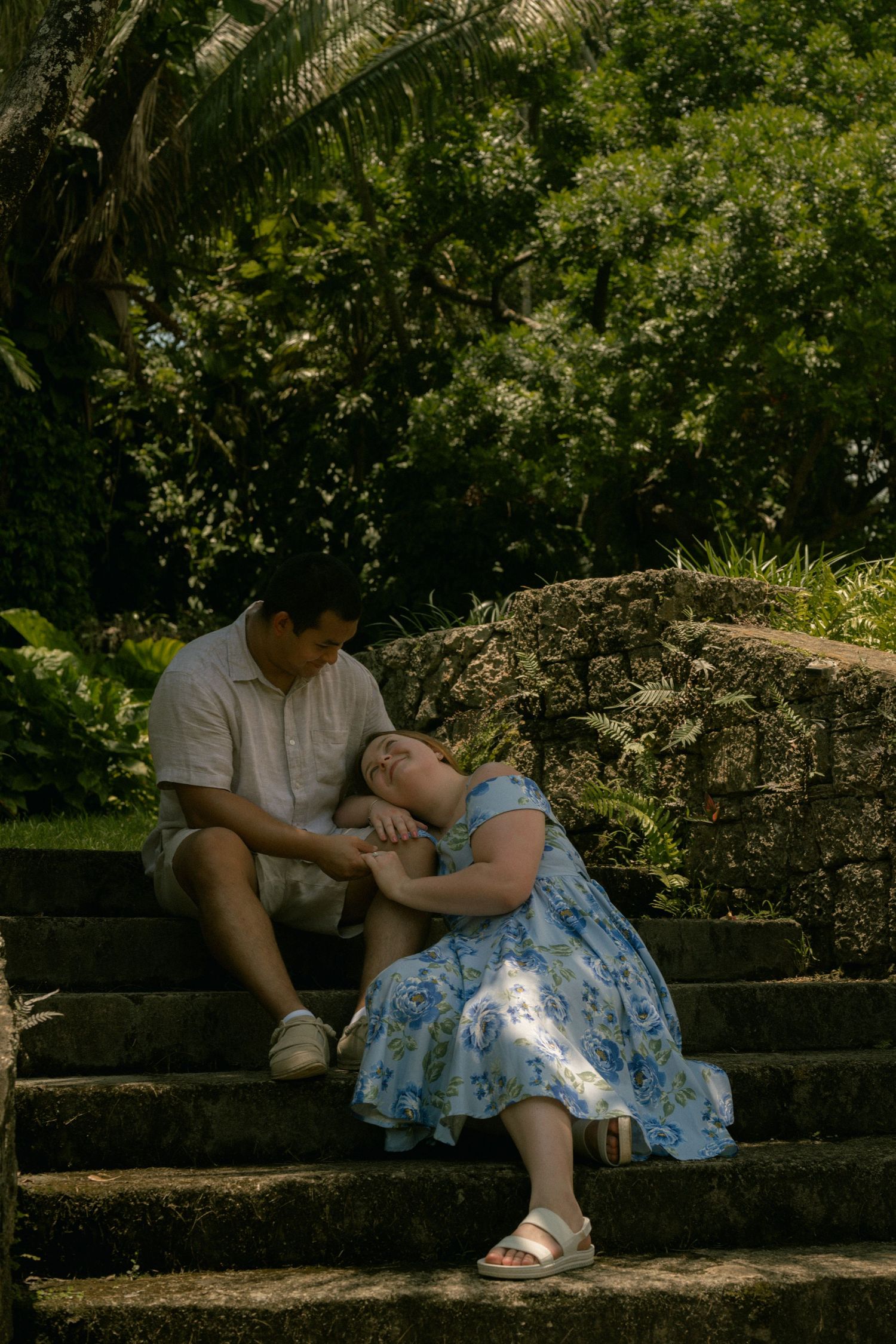 Couple sitting on stone steps in a lush garden, woman in blue floral dress leaning on mans shoulder.