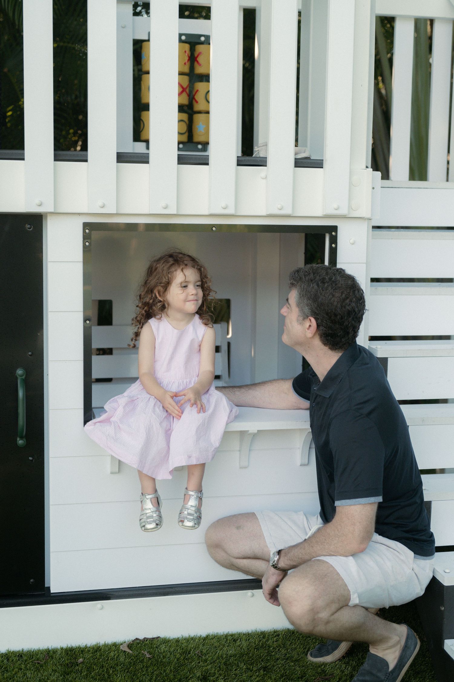 A young girl in a pink dress sits on the window ledge of a white wooden playhouse while a man crouches on the grass smiling at her.