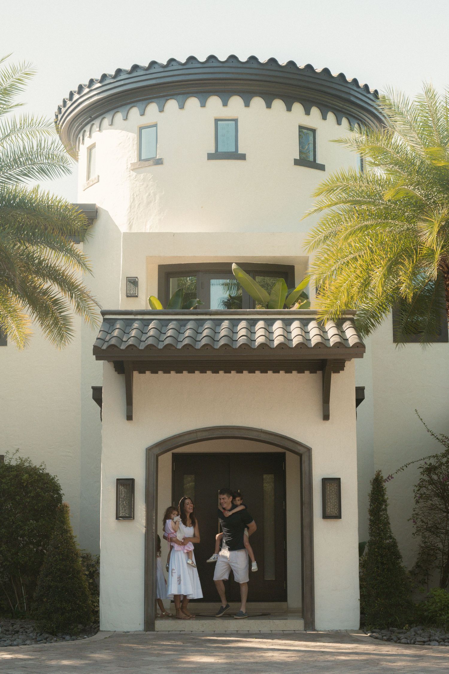 Family of four stands in the arched entryway of a cream stucco home with a round tower, tile roof, and palm trees.