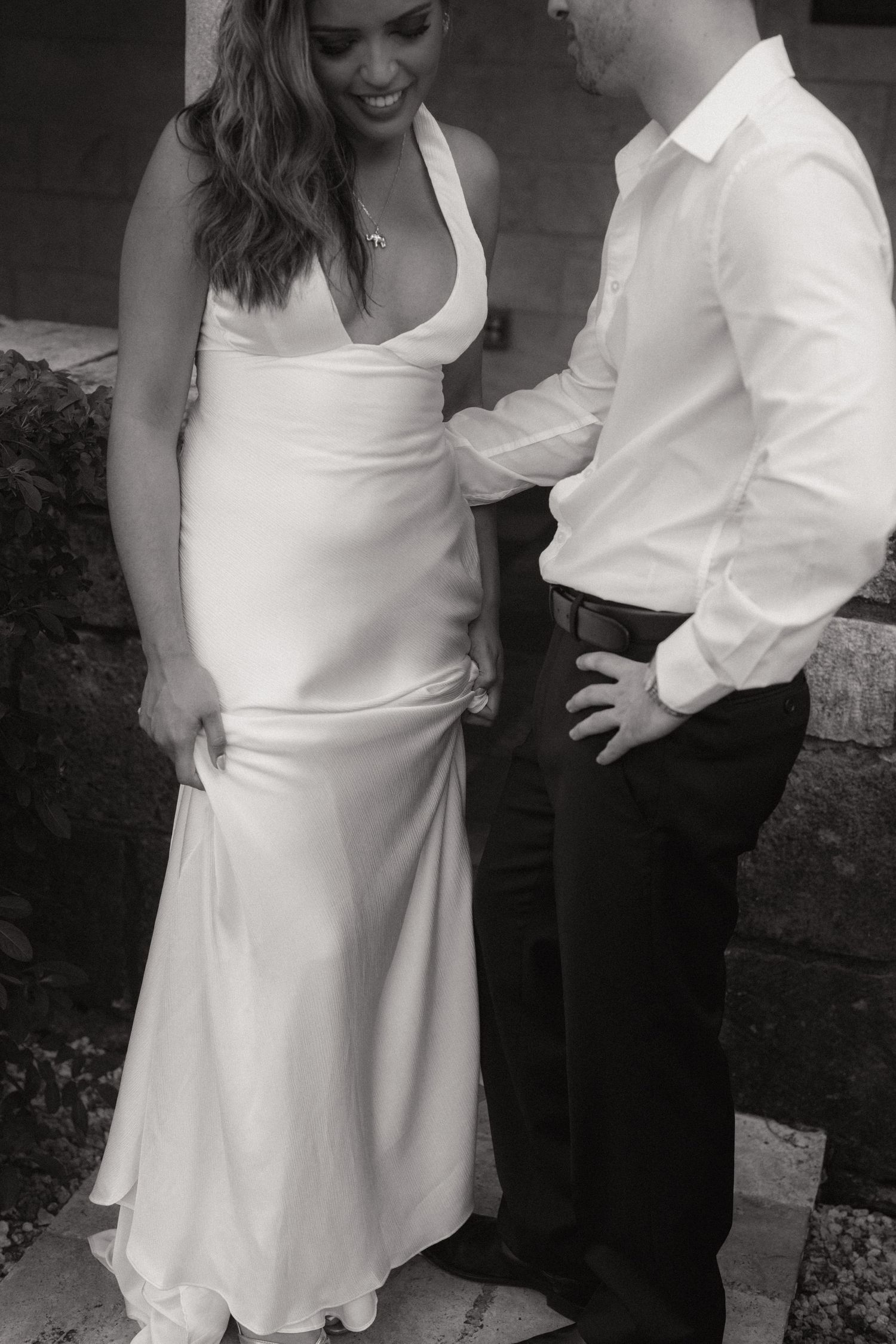 Black and white portrait of a bride in a satin gown lifting her skirt hem while her groom in a white shirt stands beside her.