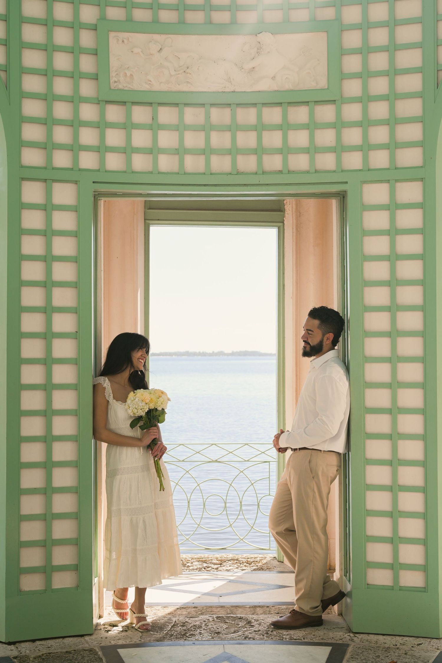 People in white clothing look through green latticed doors at a waterfront view in a beach house setting.