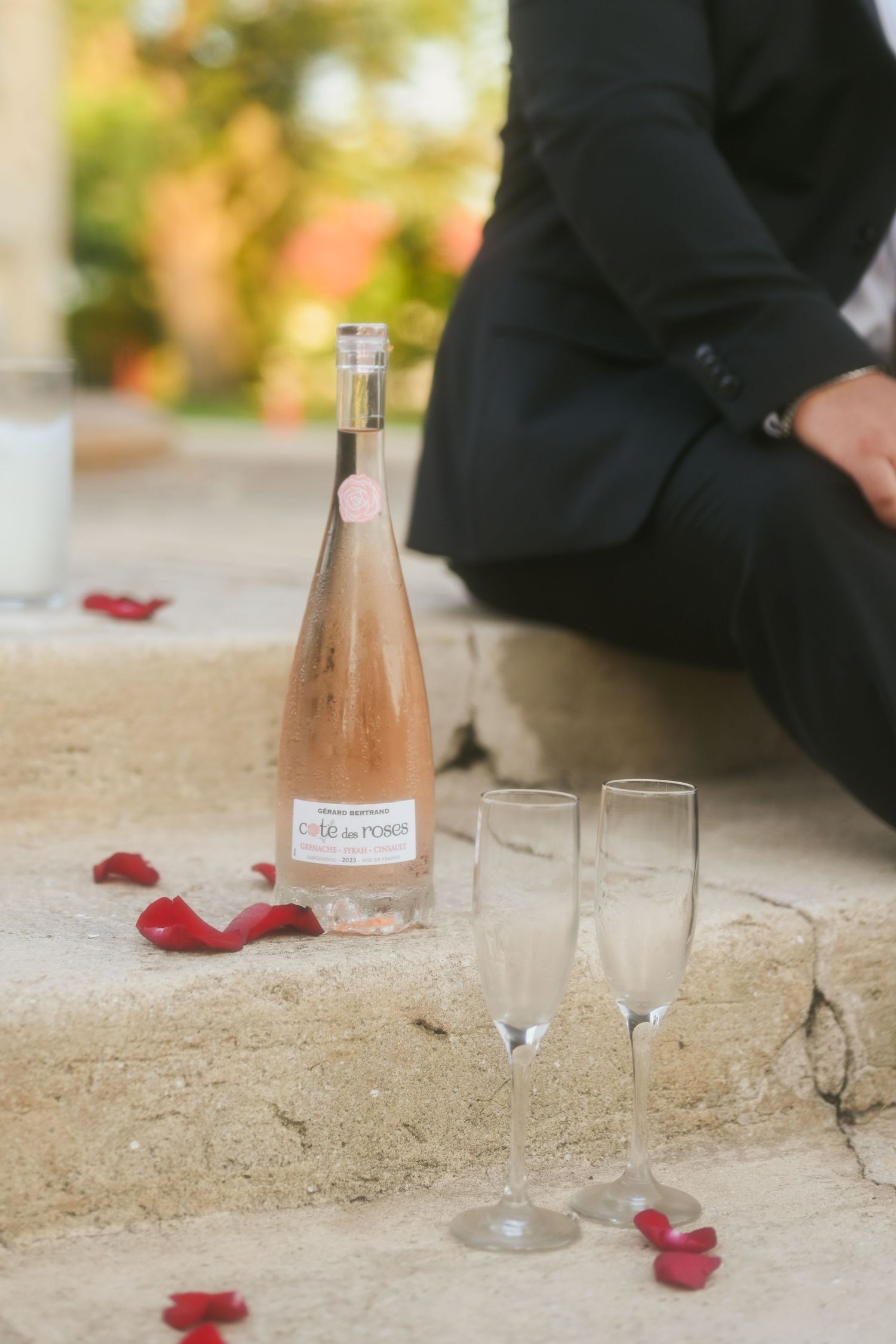 Pink champagne bottle and glasses arranged with scattered rose petals on stone surface.