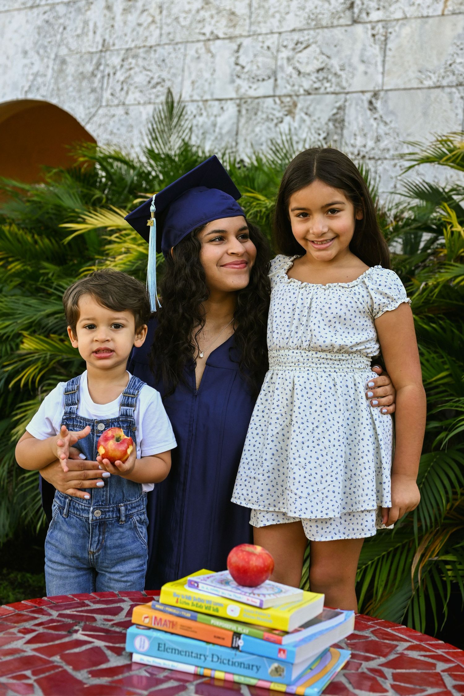Graduate in navy cap and gown poses with children and educational books outdoors.