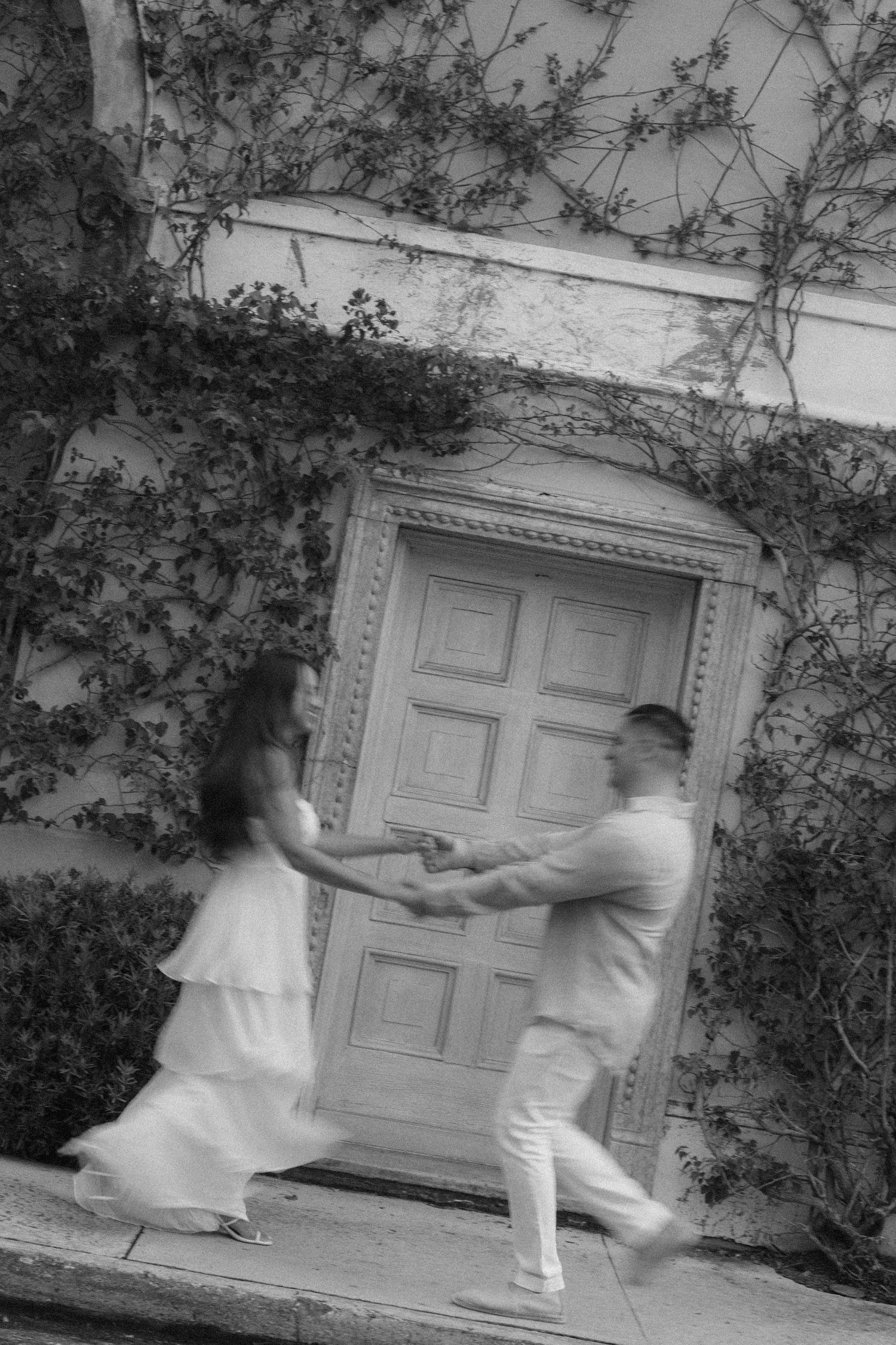 Black and white photo of a couple dancing joyfully in front of an ornate door covered in ivy vines.