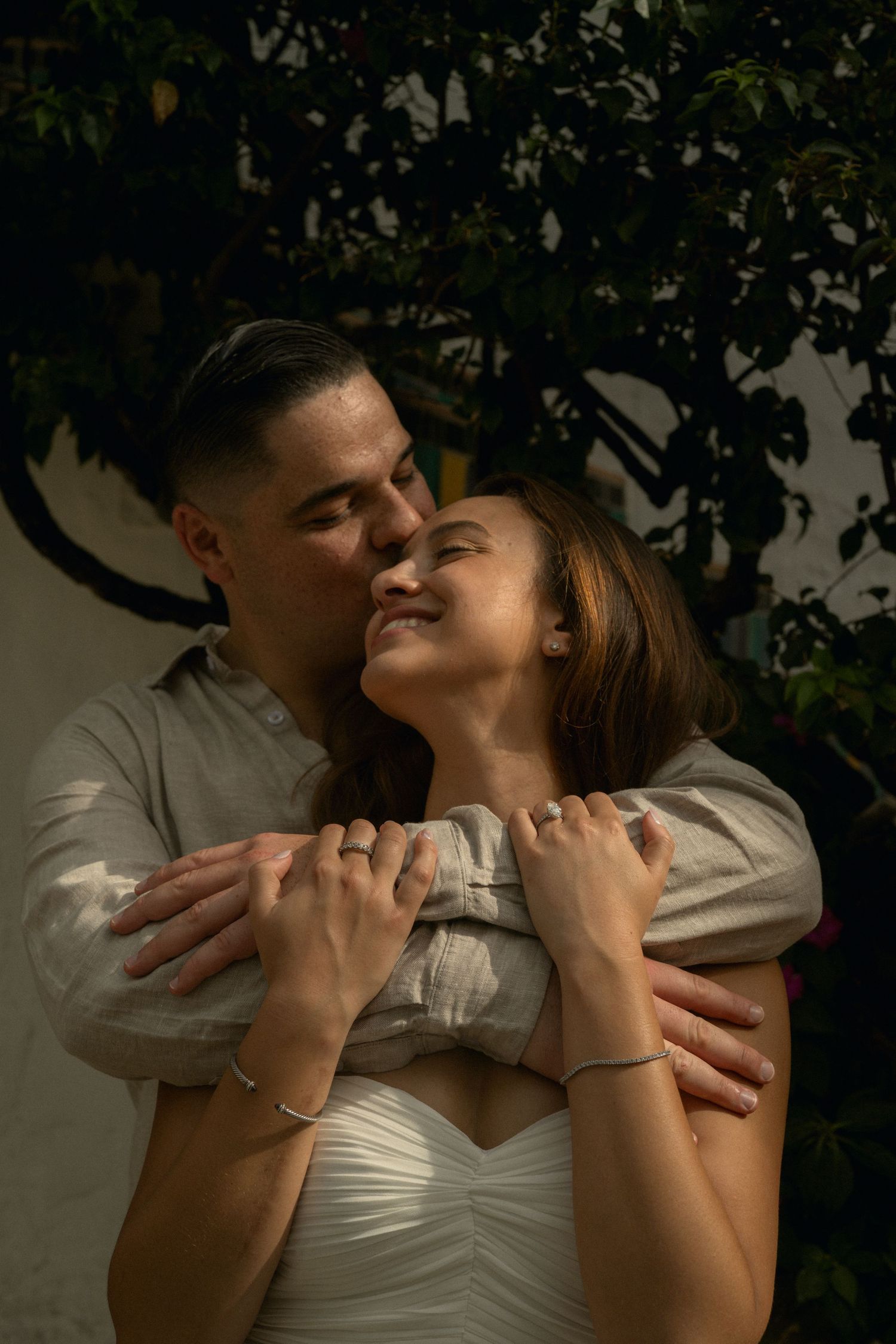 A man tenderly embraces a smiling woman from behind near a leafy green tree outdoors.