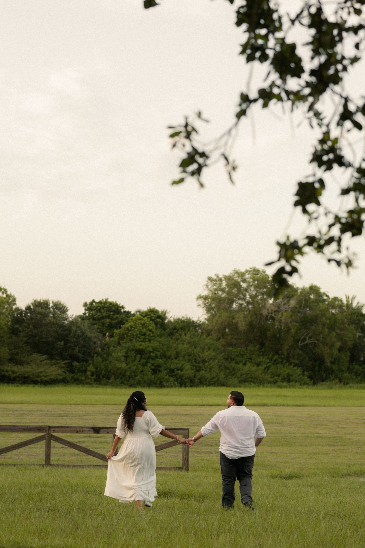 Couple holding hands walking through green field toward wooden fence, woman in white dress, framed by tree branches.