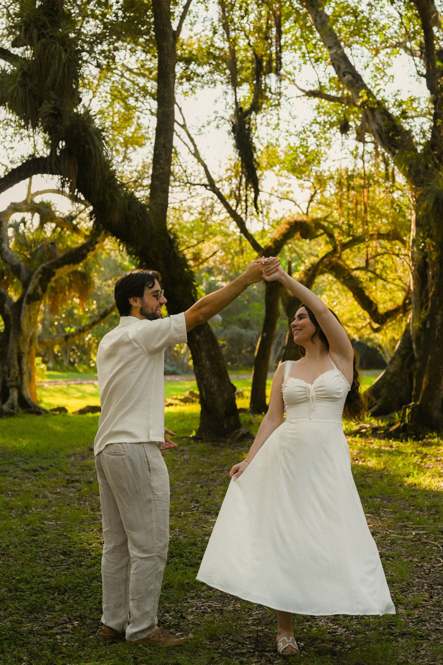 A couple in white attire dances playfully among trees in a sunlit outdoor setting during their wedding photoshoot.
