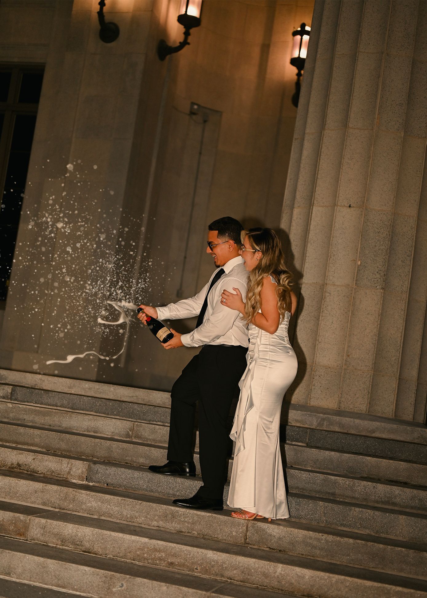 Wedding couple celebrating with champagne on stone steps at night.