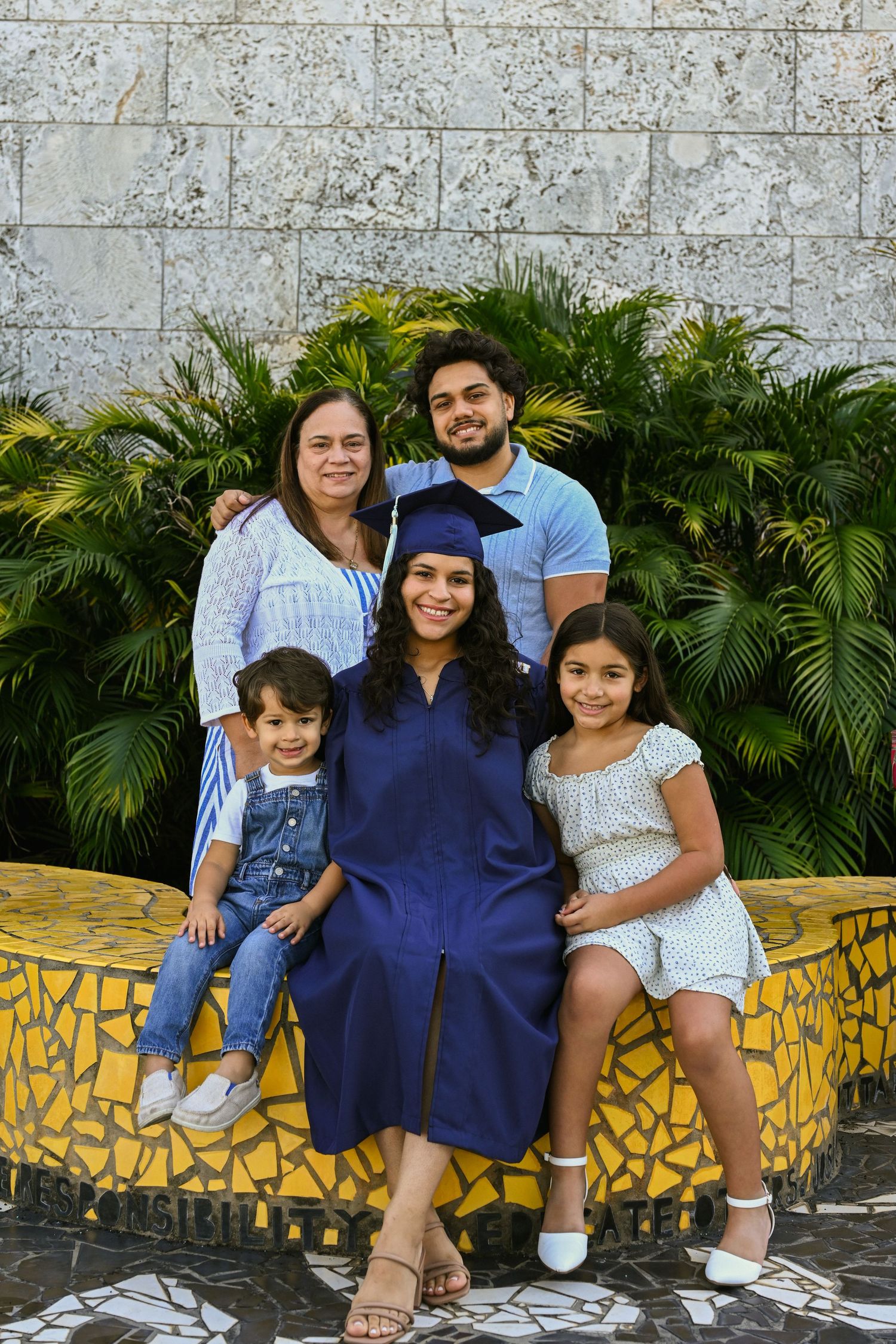 Family graduation portrait with palm plants and decorative mosaic wall background.