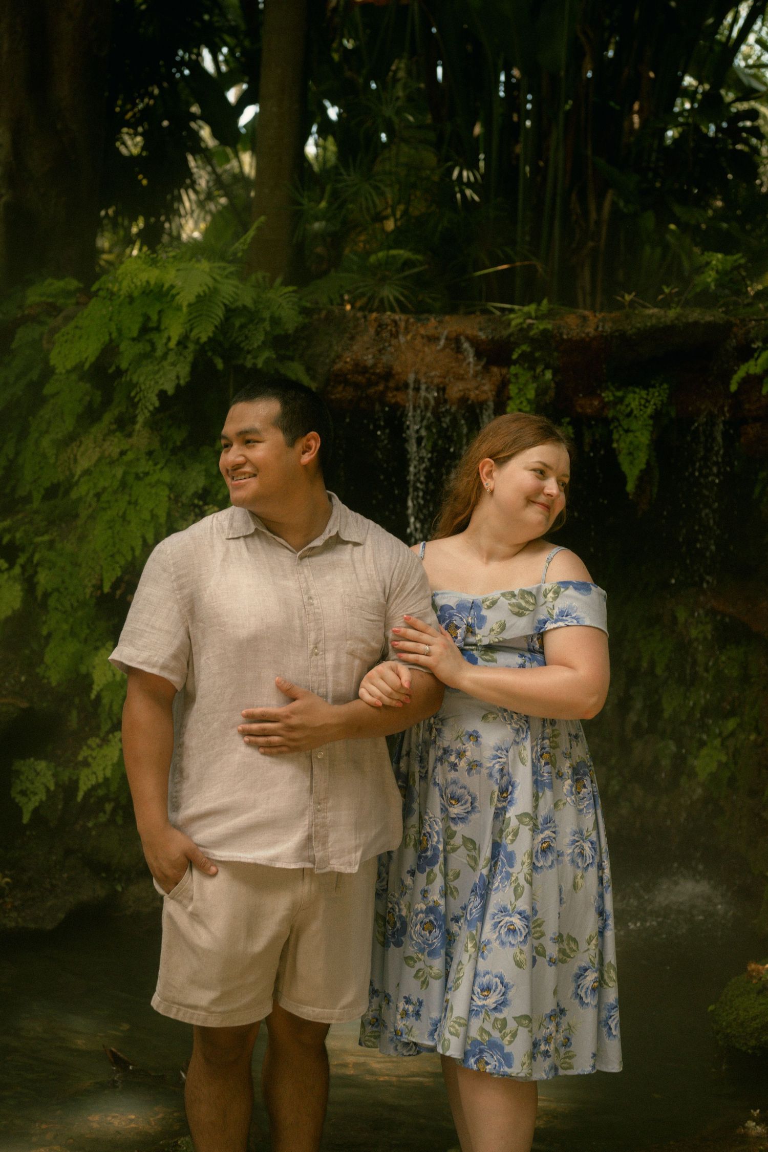 A smiling couple walks arm in arm near a waterfall surrounded by tropical greenery, wearing casual summer outfits.