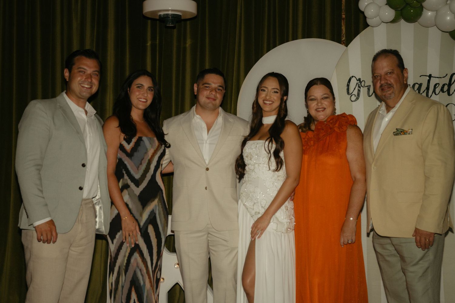 Group of six people posing together at a graduation celebration party with decorated backdrop.