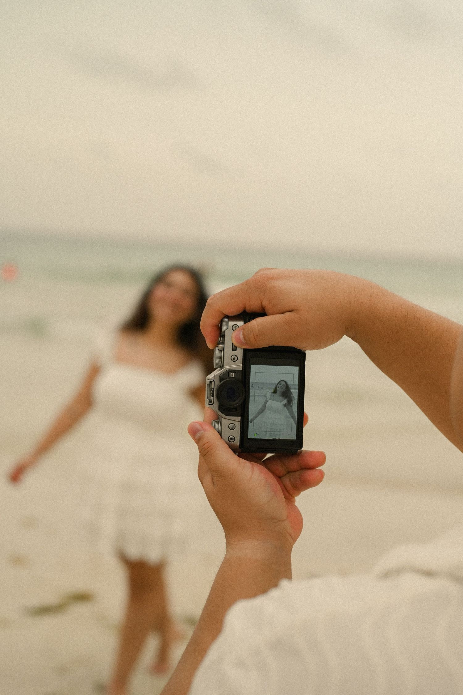 A person holds a digital camera at the beach to take photos of distant figures in soft, dreamy lighting.