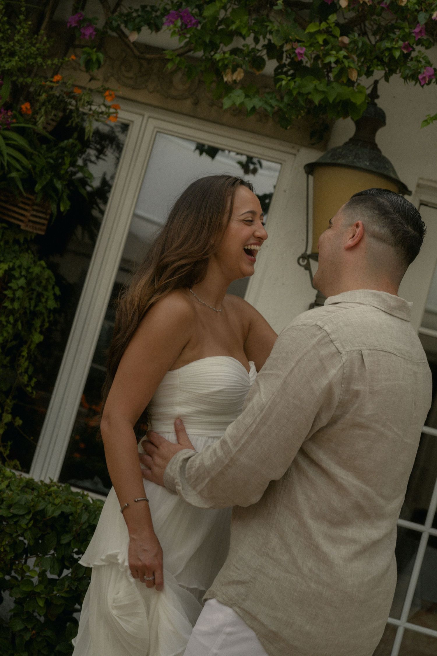 A laughing bride in a white strapless gown shares a joyful moment with her groom in a beige linen suit outdoors.