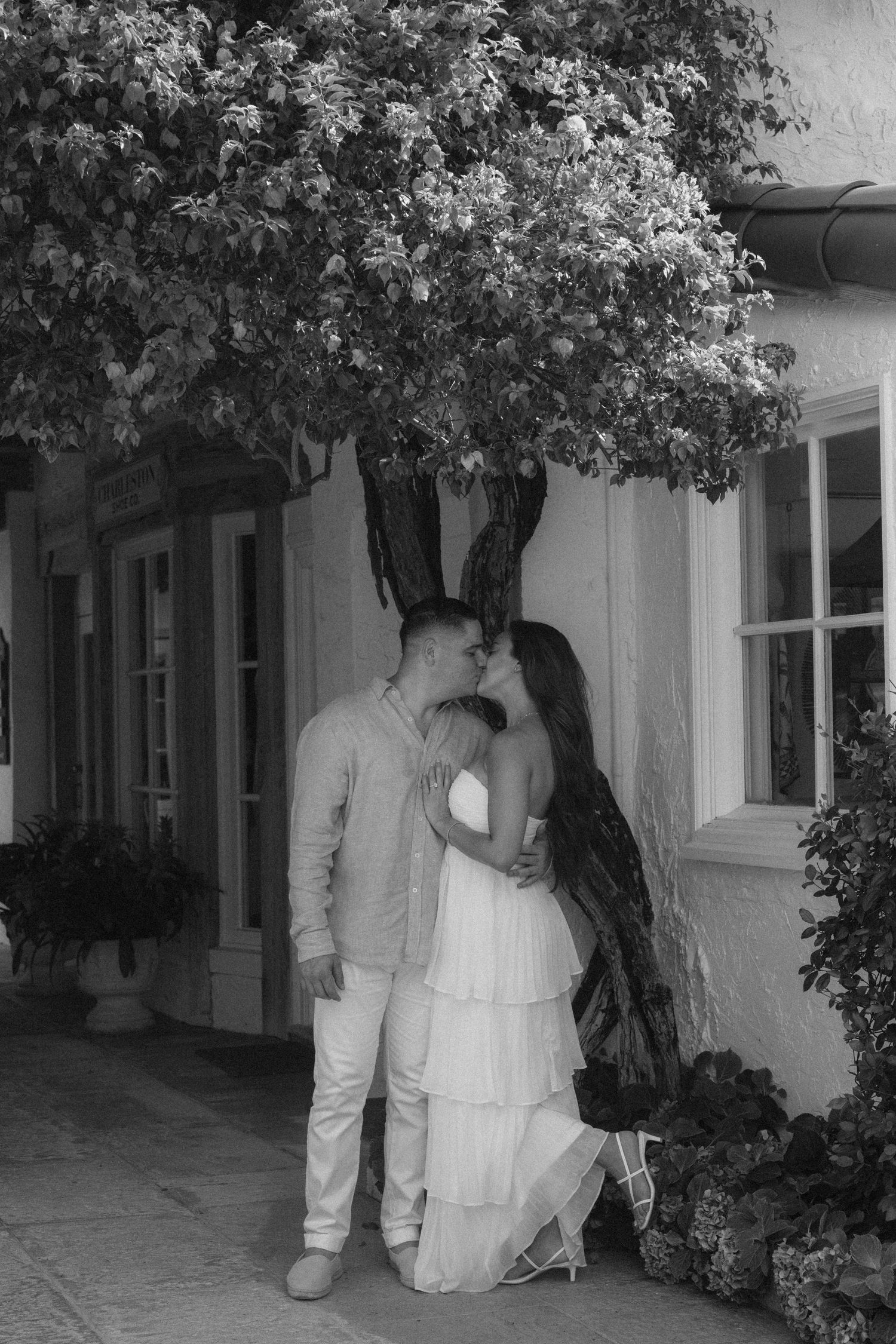 Black and white photo of couple kissing under flowering tree beside charming cottage with white windows.