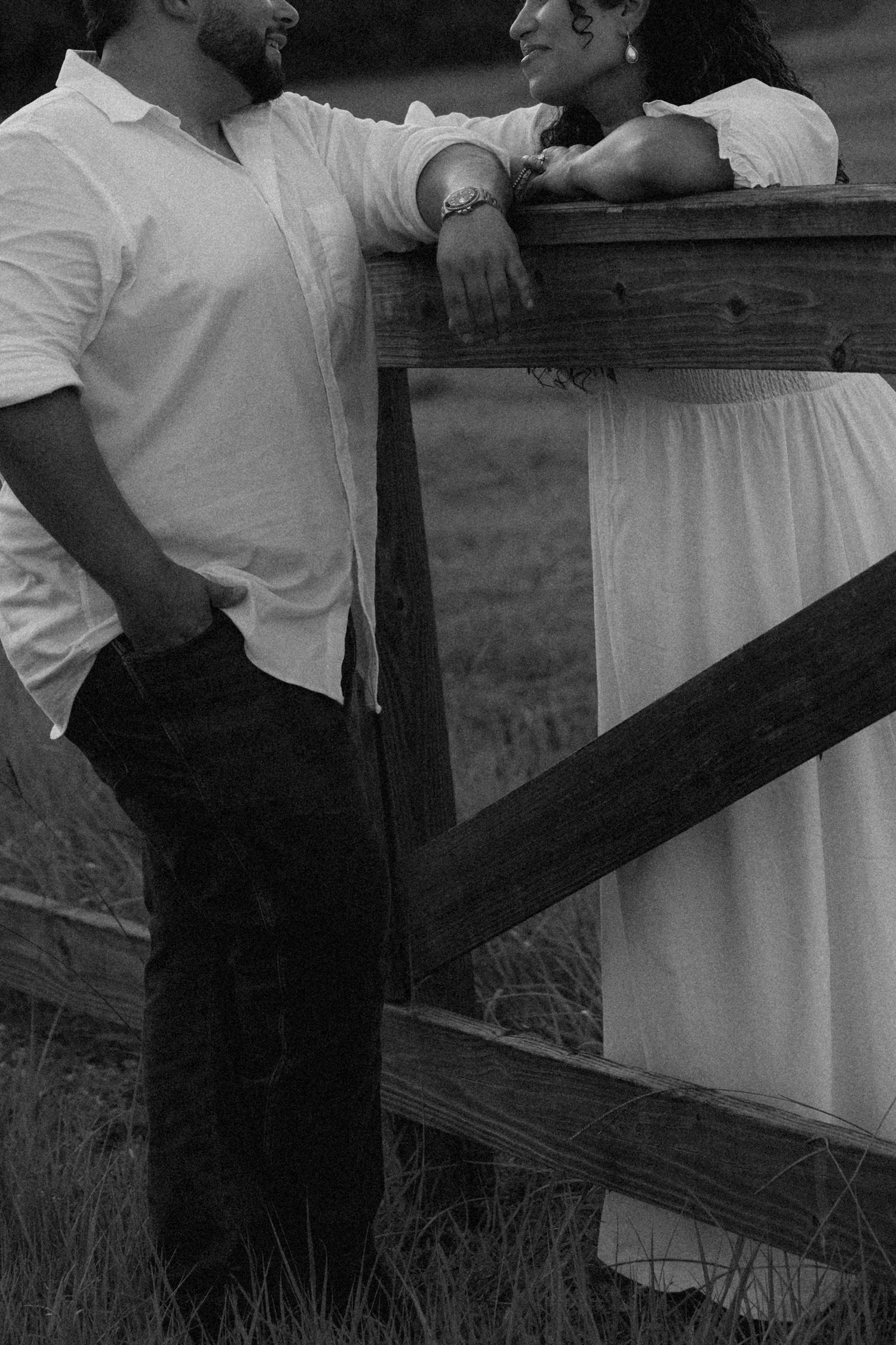 Black and white photo of a couple leaning on a rustic wooden fence in a field, sharing a romantic moment.