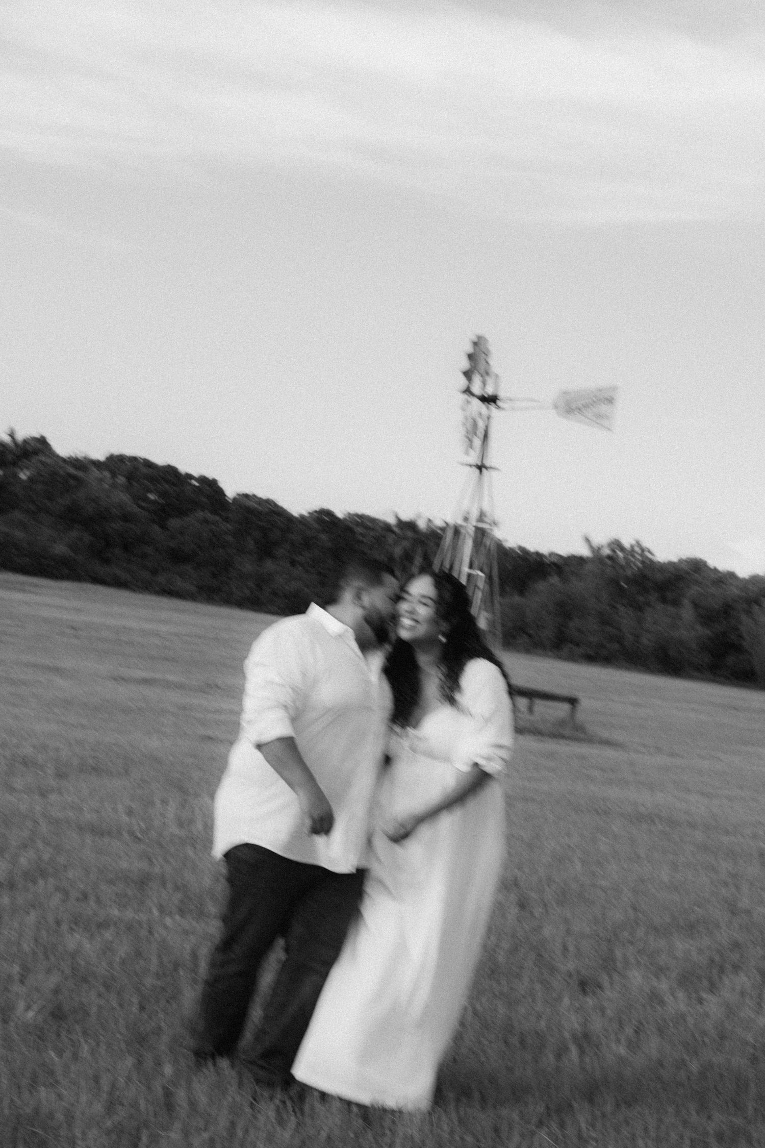Black and white photo of a couple embracing in a field with a windmill backdrop during a romantic outdoor maternity session.