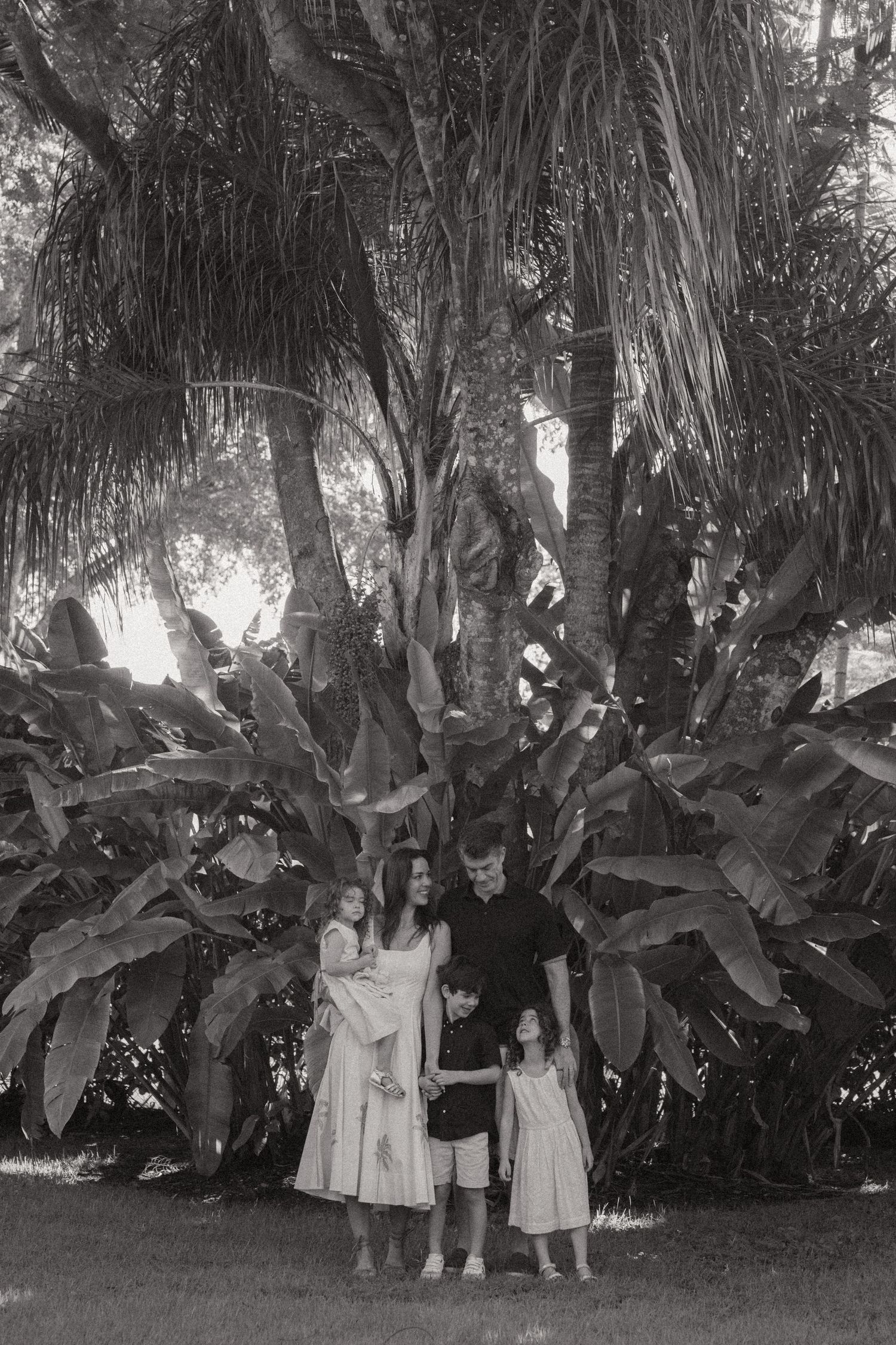 Black and white family portrait of two parents and three children standing together beneath tall palm trees and large tropical leaves.