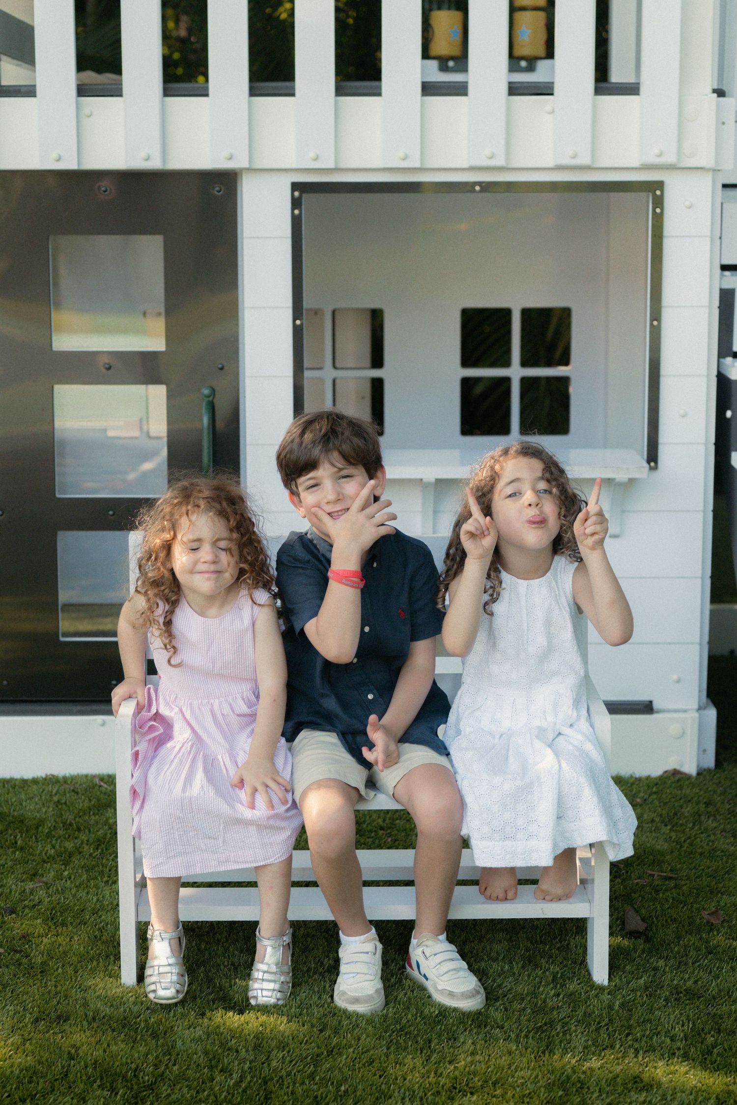Three smiling children sit together on a white bench in front of a white playhouse on green grass, making playful hand gestures.