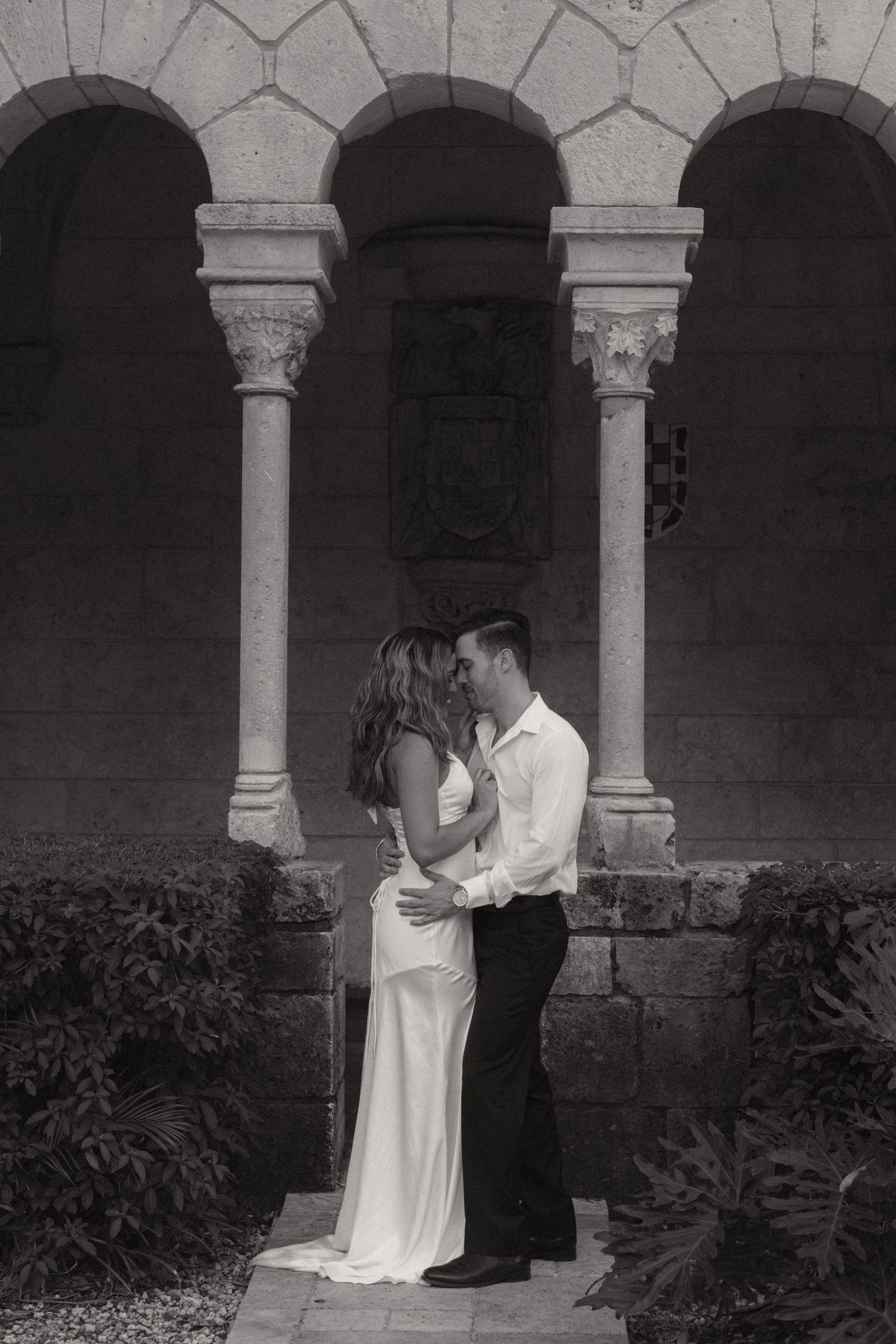 Black and white engagement photo of a couple embracing beneath stone arches and columns in a historic courtyard garden.